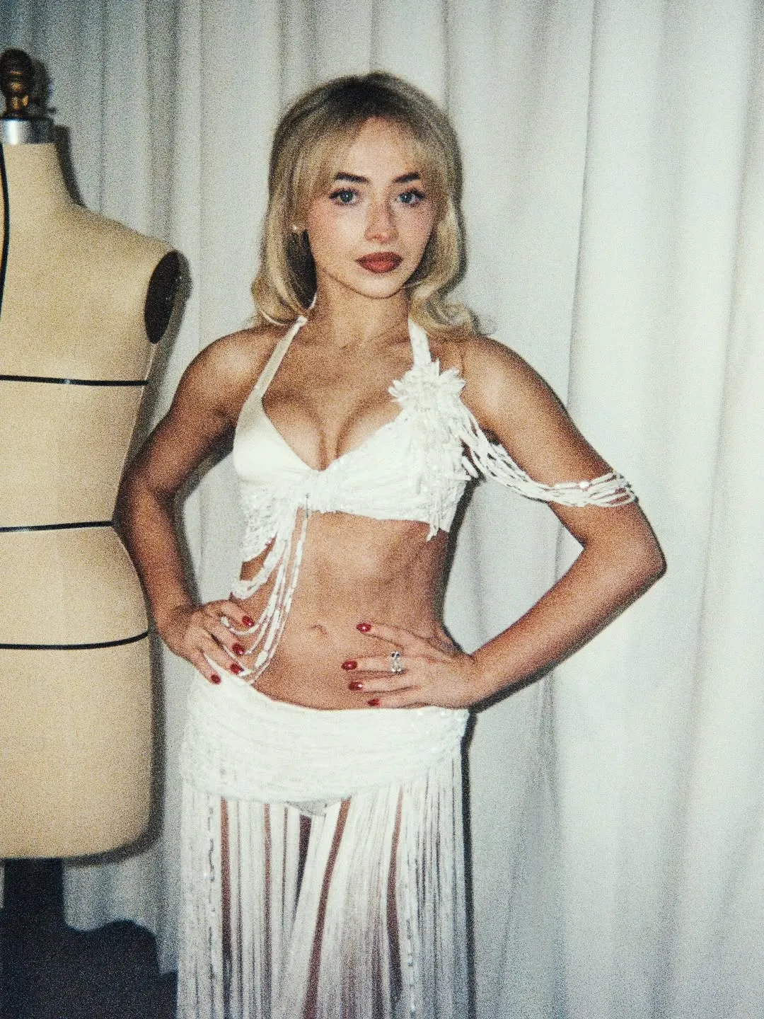 Woman in a white fringe outfit posing with hands on hips, mannequin visible in the background.