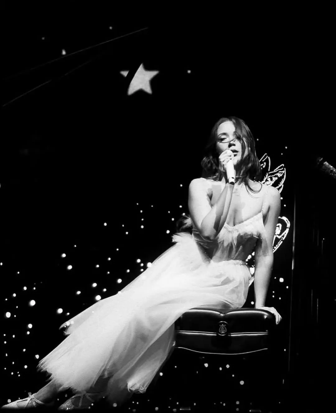 Female singer in a white dress performs on a dark stage, holding a microphone amid starry lights.