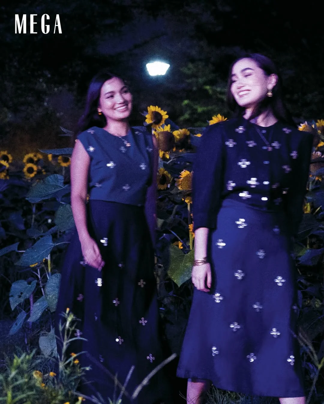 Two women in matching navy outfits with white embroidery stand among sunflowers at night, smiling. A MEGA cover vibe.