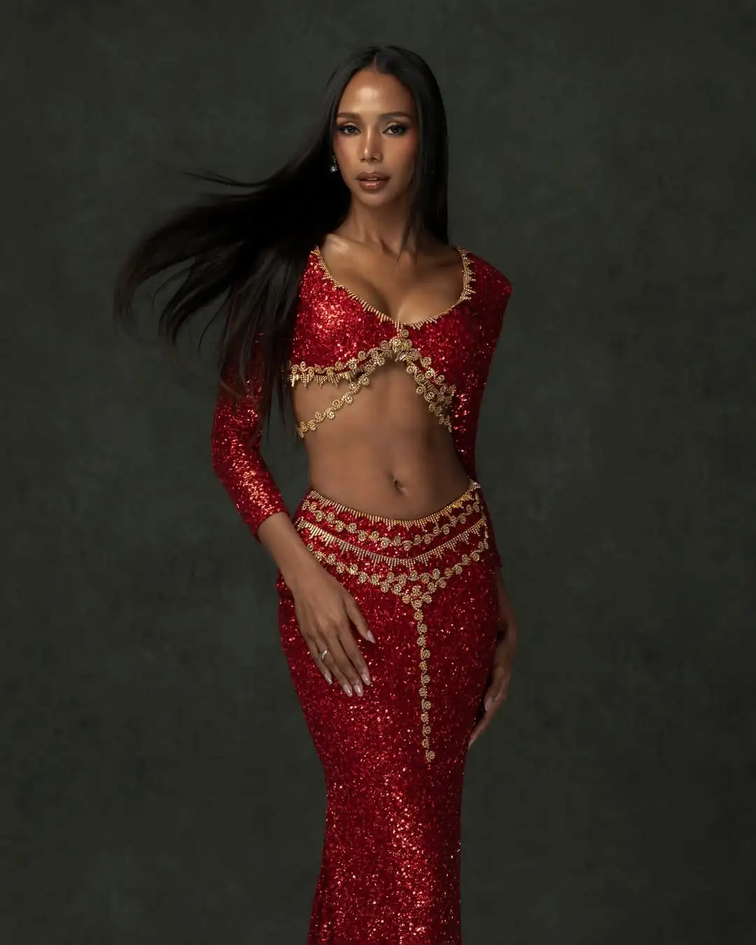 Portrait of a woman in a red sequin belly-dance outfit with gold trim posing against a dark backdrop.