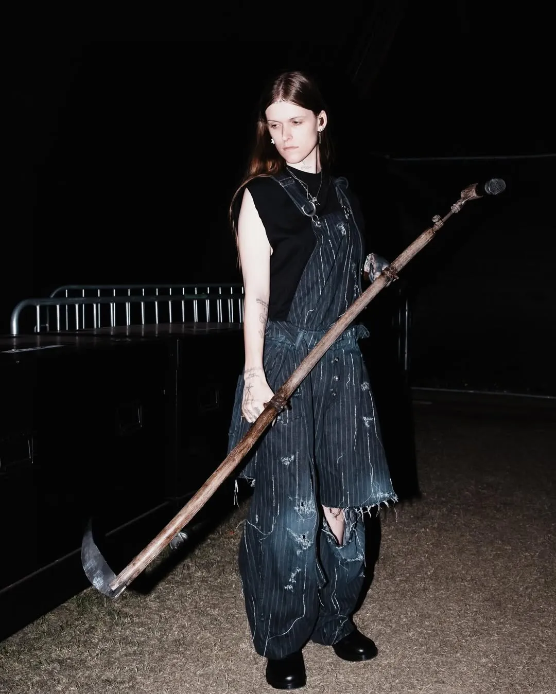 Person with long hair wearing distressed denim overalls and a black sleeveless top, holding a long wooden axe-like prop outdoors at night.