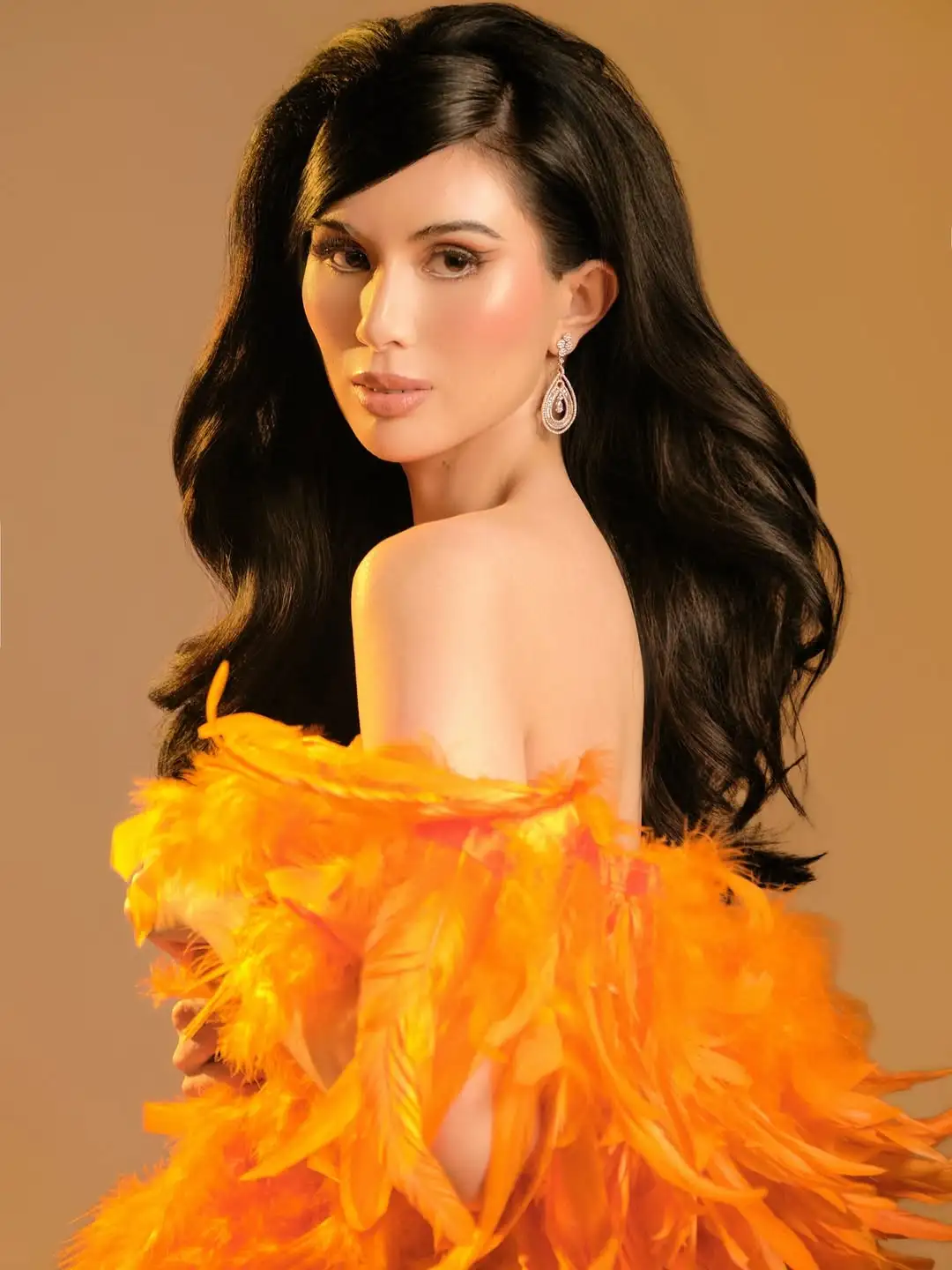 Portrait of a woman with long dark hair wearing an off-shoulder orange feathered dress and teardrop earrings against a warm backdrop.