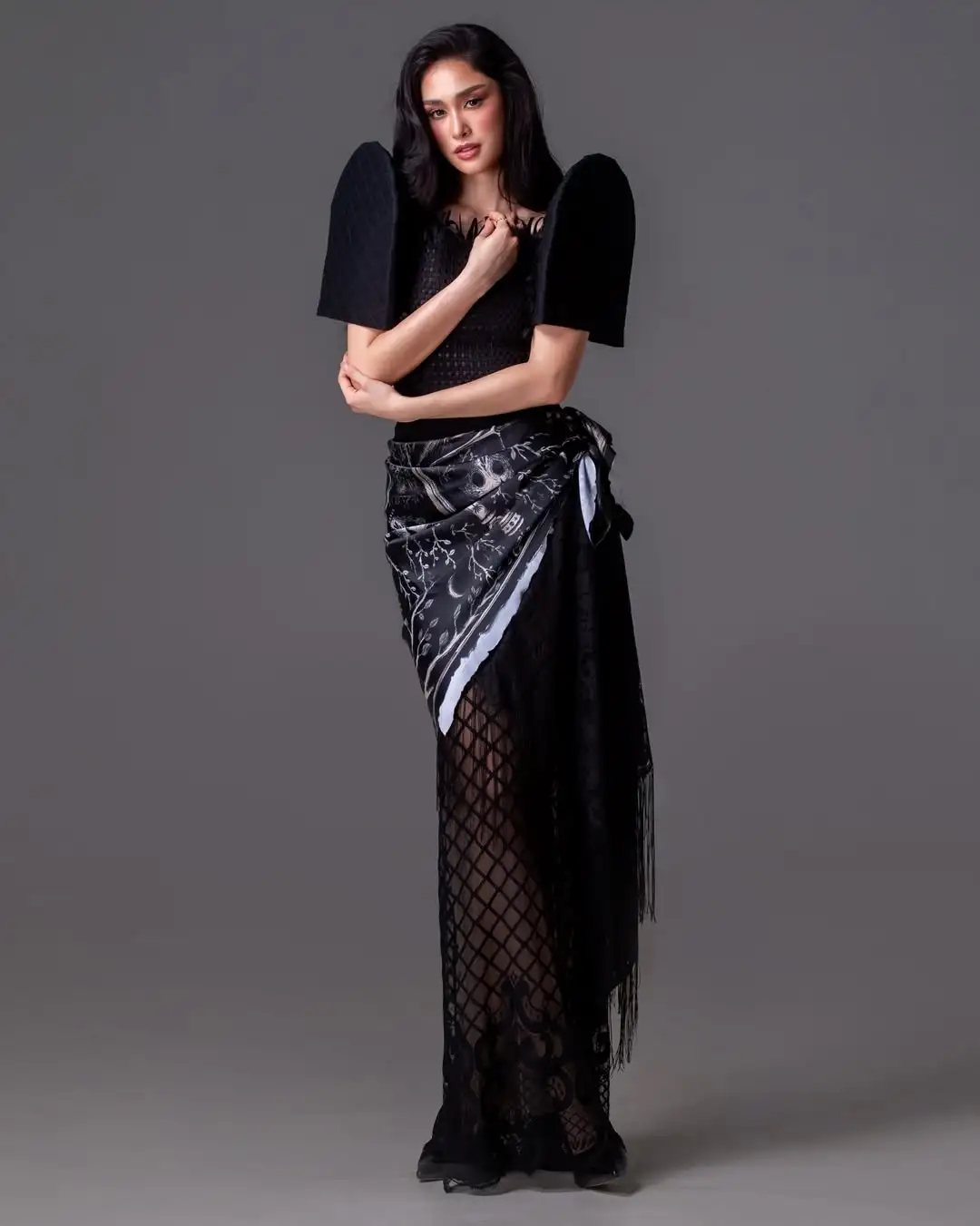 Model wearing a black haute-couture outfit with large wing sleeves, a patterned wrap skirt, and fishnet tights, posing.
