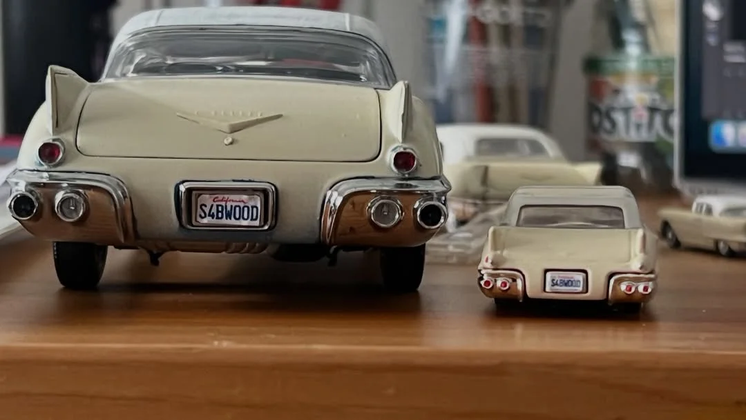 Two vintage-style beige toy cars seen from the rear on a wooden surface, chrome bumpers and tail lights visible.