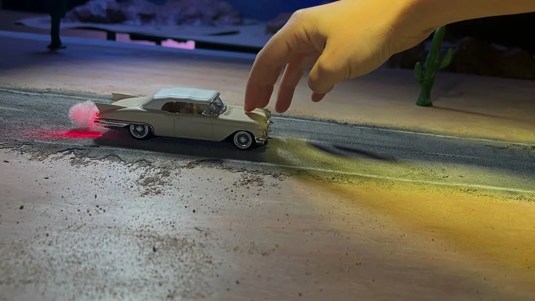 A small vintage toy car on a miniature road, a large hand hovering above it as if about to push or stop the car.