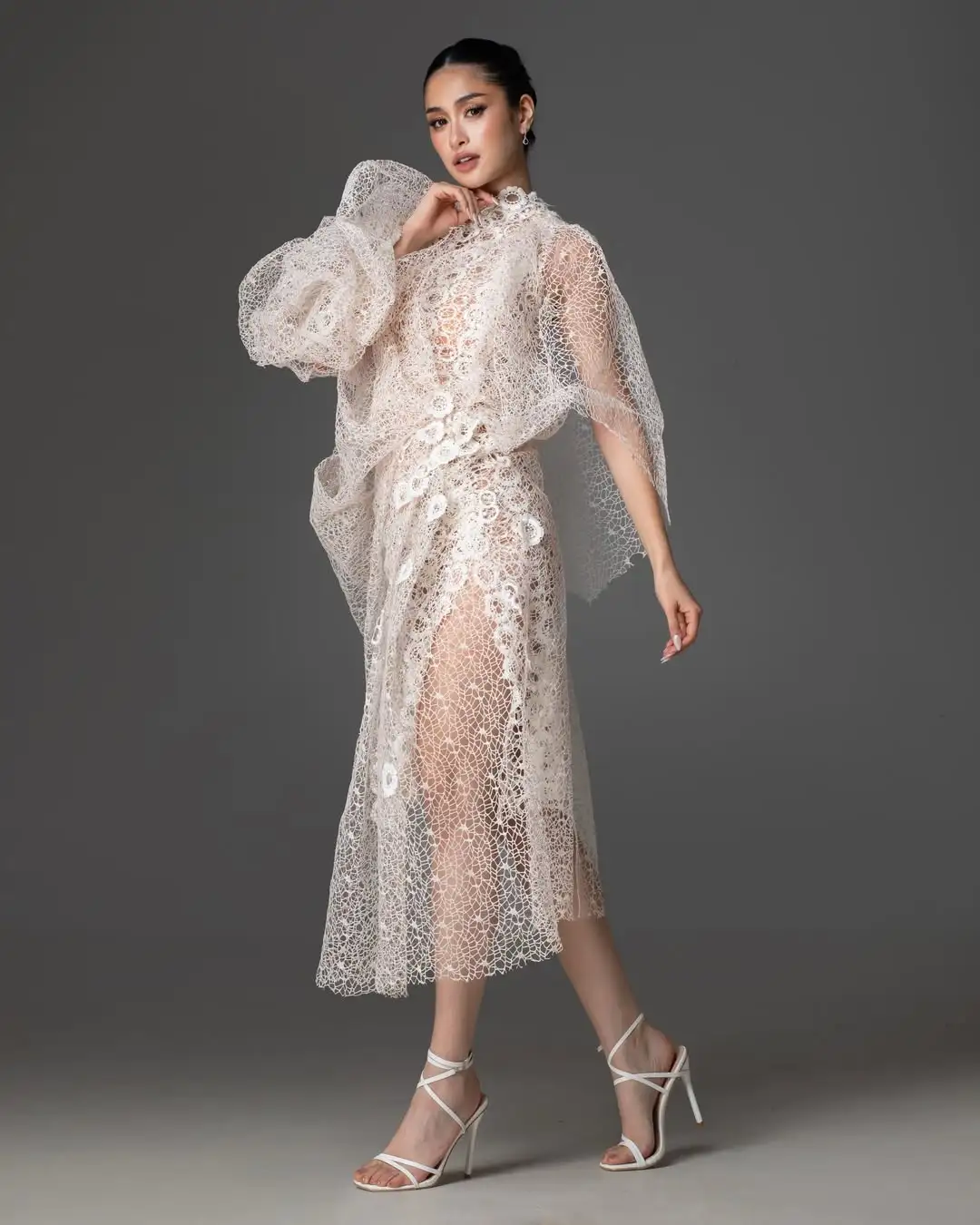 Model in a cream lace midi dress with sheer overlay and voluminous sleeves, wearing white strappy heels.