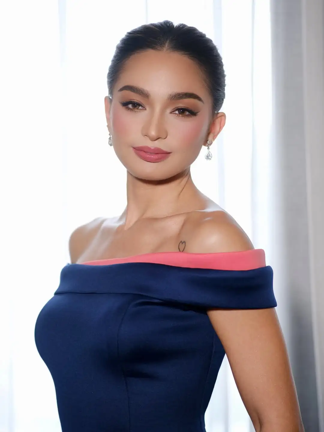 Portrait of a woman with dark hair in a sleek updo, wearing an off‑the‑shoulder navy dress with a pink neckline and dangling earrings, looking confidently at the camera.