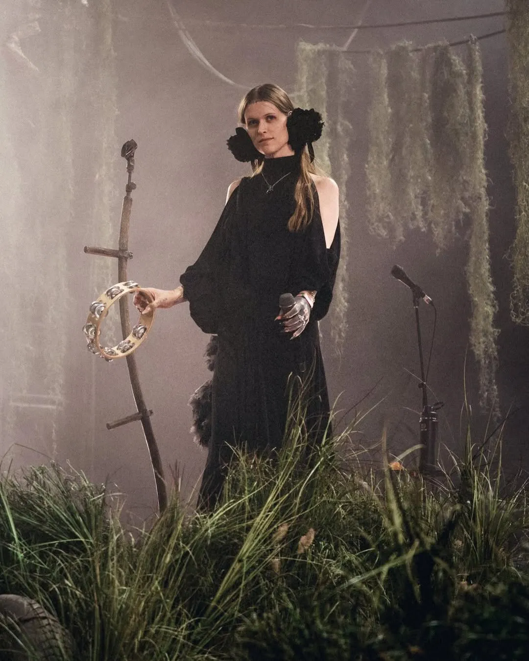 Female performer in a black dress on a smoky stage, holding a tambourine with greenery in the foreground and a microphone nearby.]','Female singer wearing an off-shoulder black gown and large black flower earrings, holding a tambourine on a moody stage.