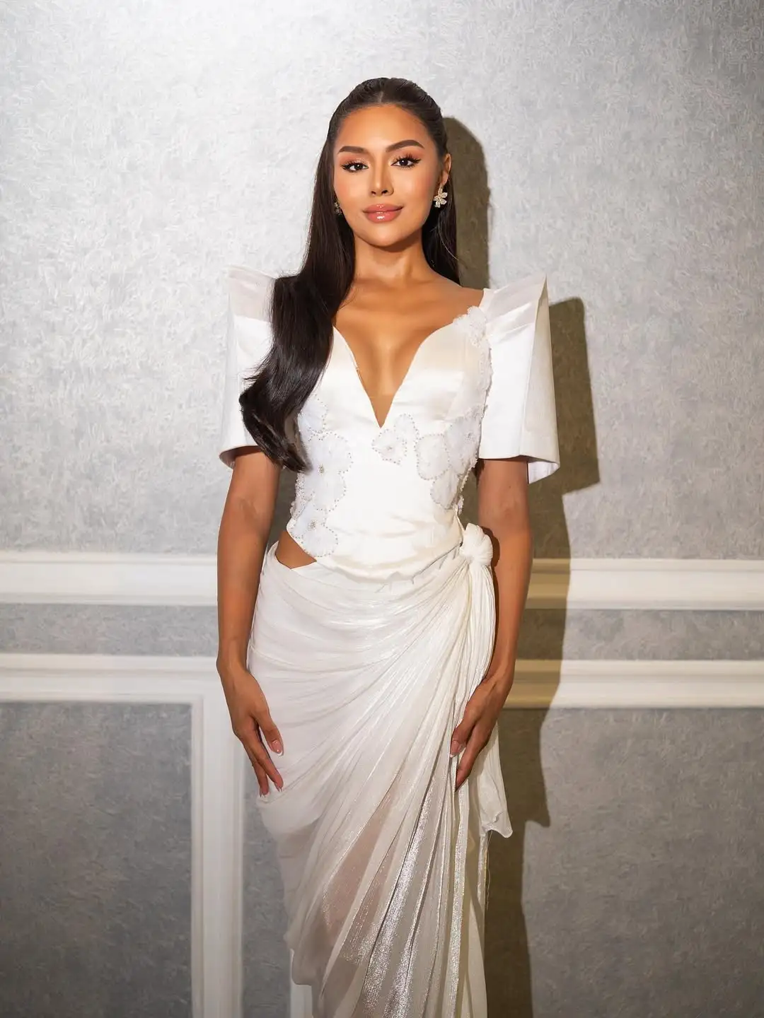 Woman in a white, elegant gown with a deep V neckline standing against a textured wall, hands at her sides, long dark hair styled straight.
