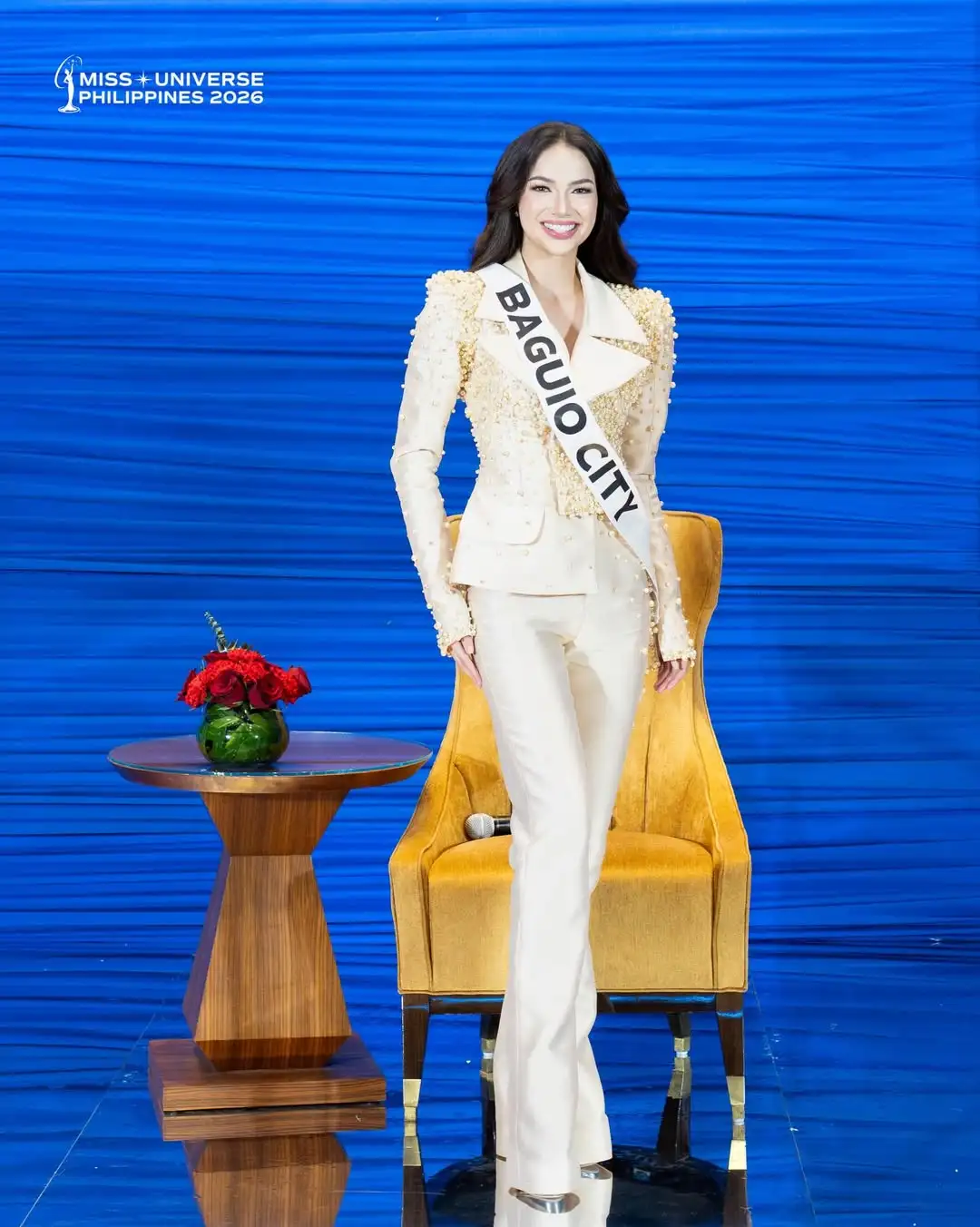 Miss Universe Philippines 2026 contestant wearing a white suit with a 'BAGUIO CITY' sash on a blue stage backdrop.