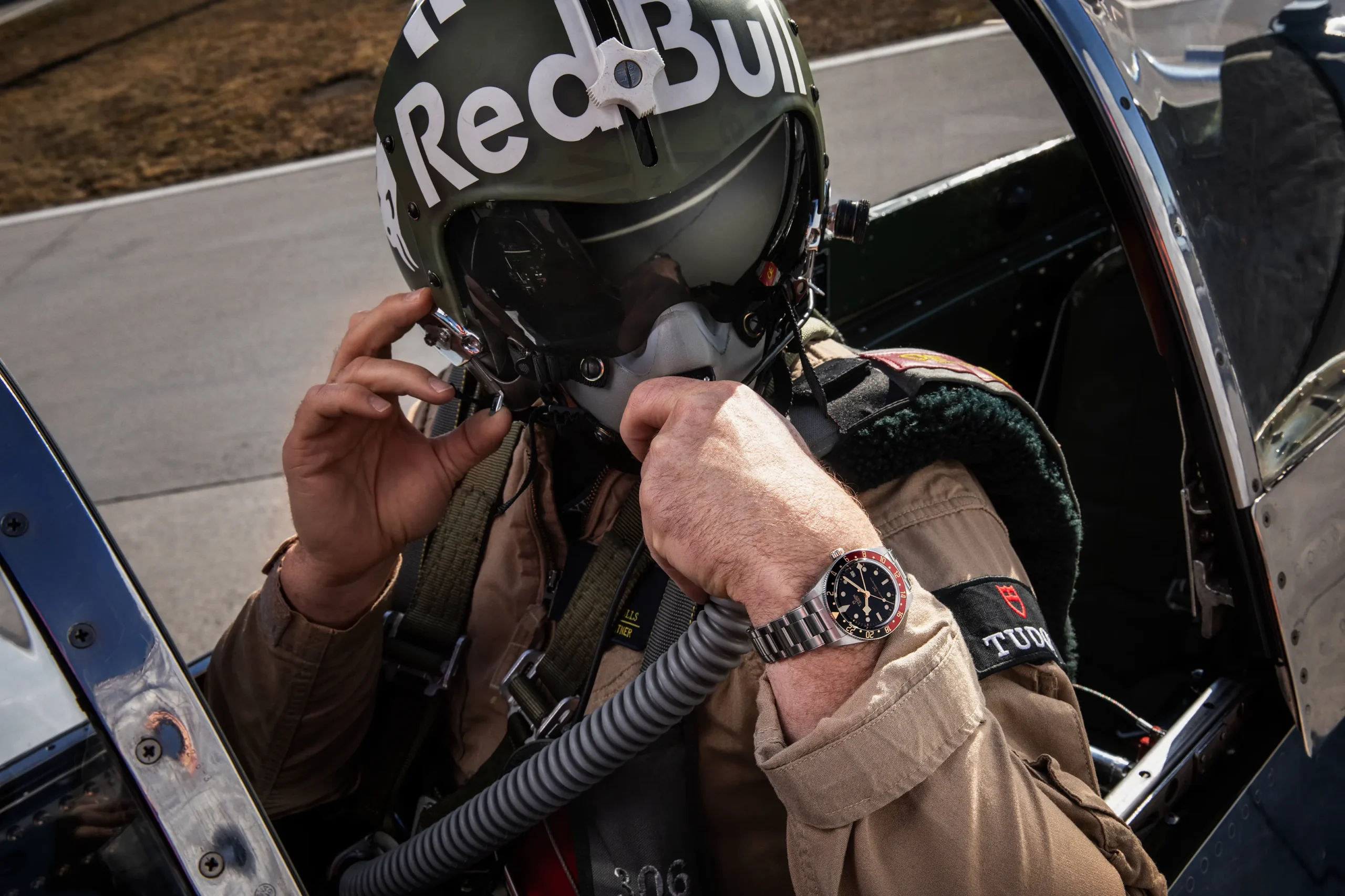 Pilot in a flight suit adjusts his helmet inside an open cockpit, wearing a Red Bull helmet with a reflective visor and a silver watch visible on his wrist