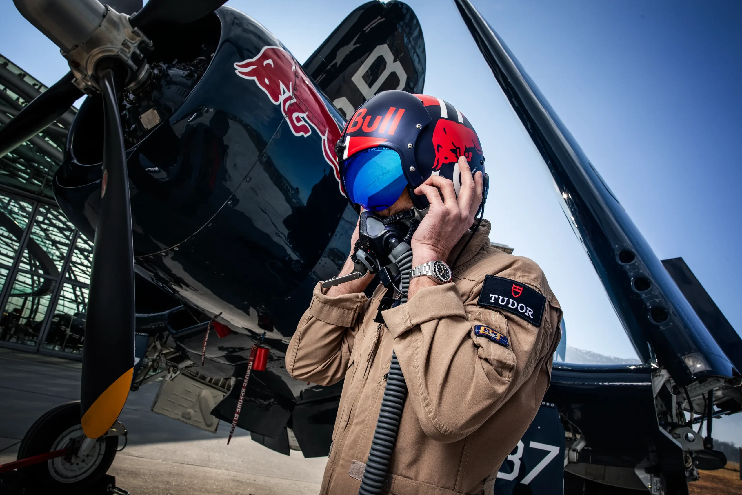 Pilot in a tan flight suit adjusts an oxygen mask beside a black propeller aircraft with Red Bull branding on the helmet helmet.