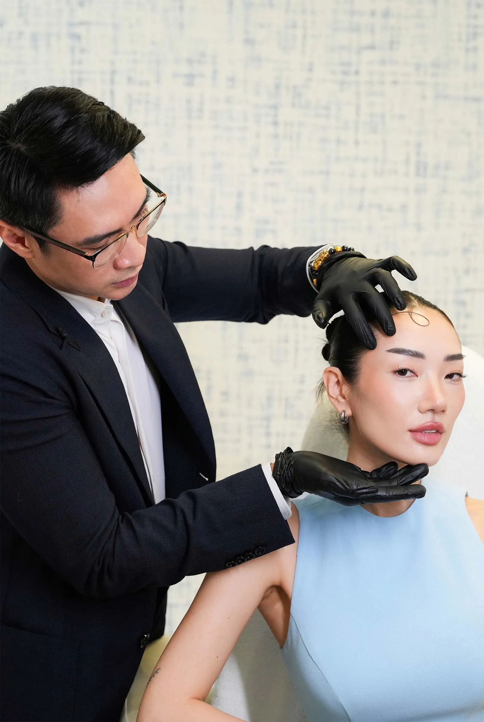 Male practitioner in a suit wearing gloves applies a cosmetic treatment to a woman's head.