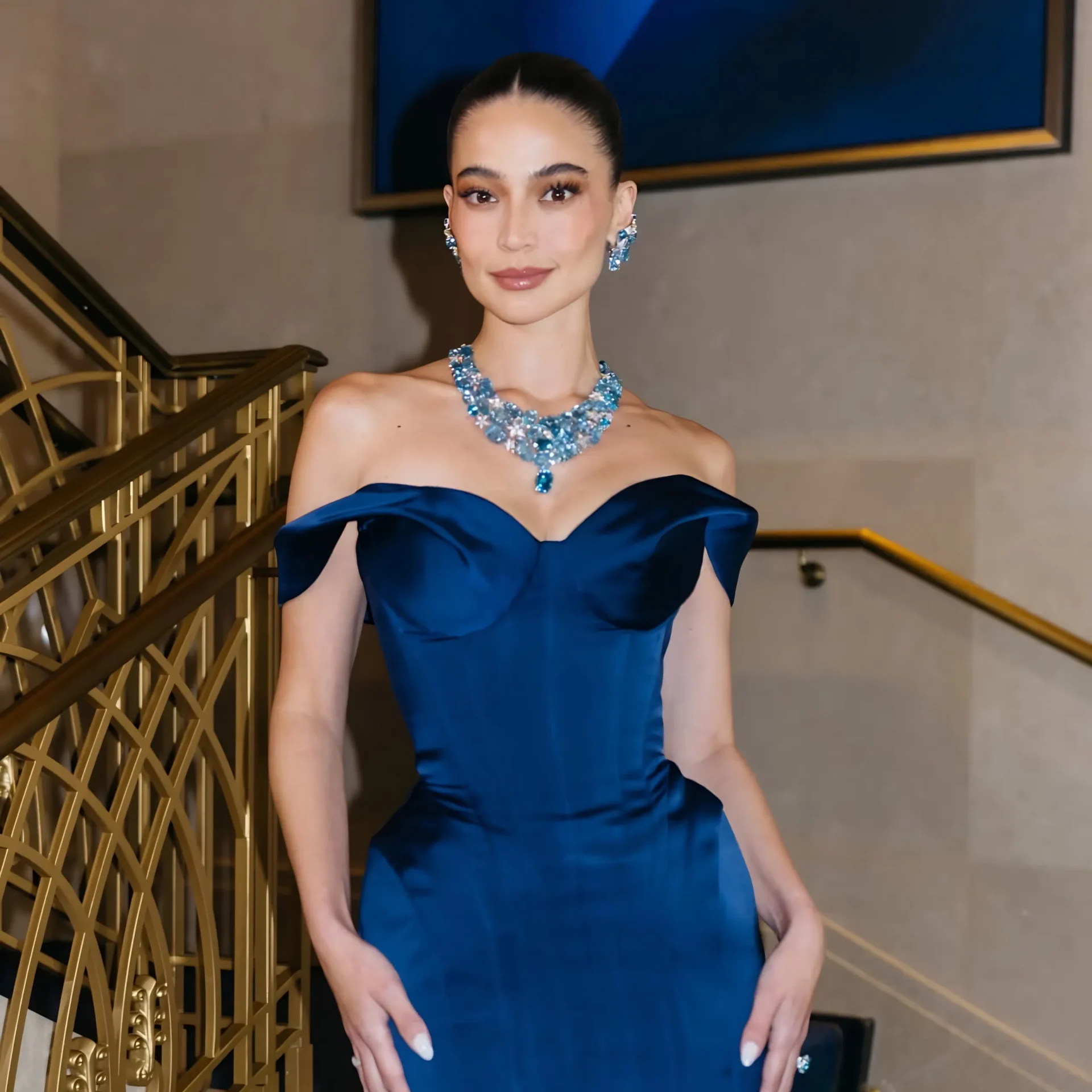 Woman in a blue off-shoulder evening gown wearing matching blue jewelry on a staircase.