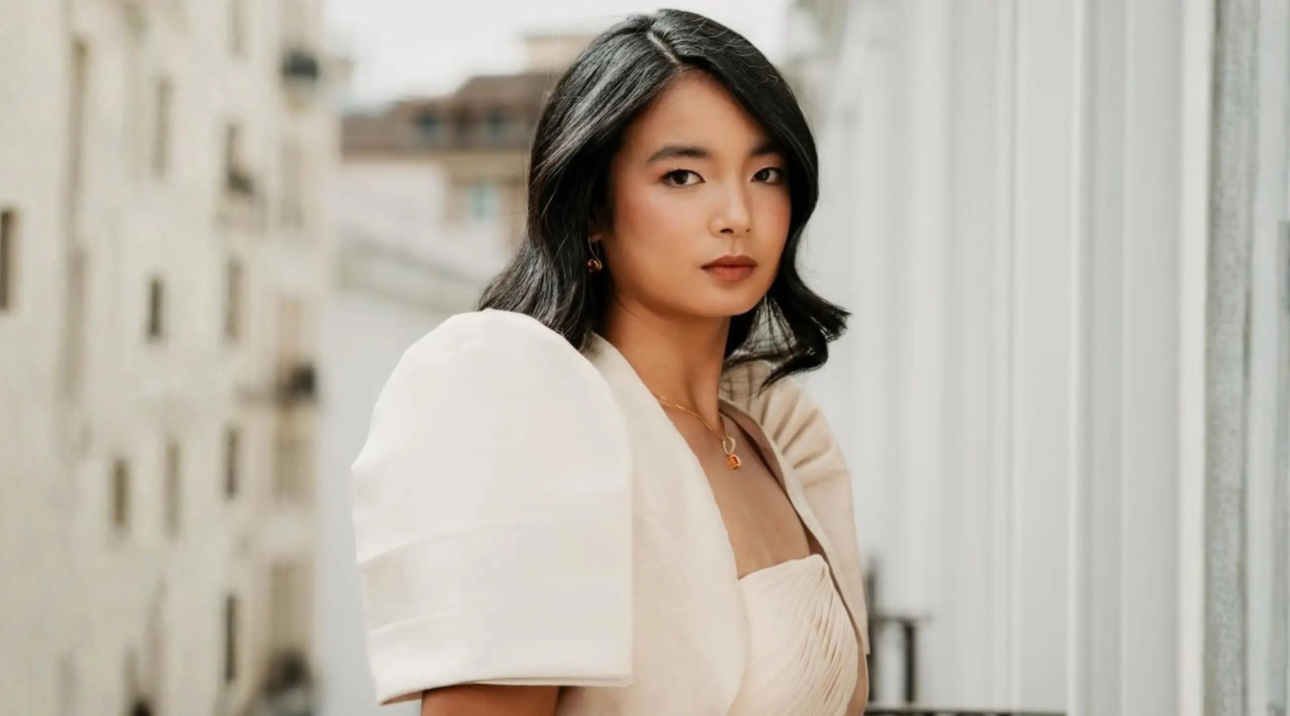 Portrait of a woman with short black hair wearing a cream blouse and gold necklace outdoors.