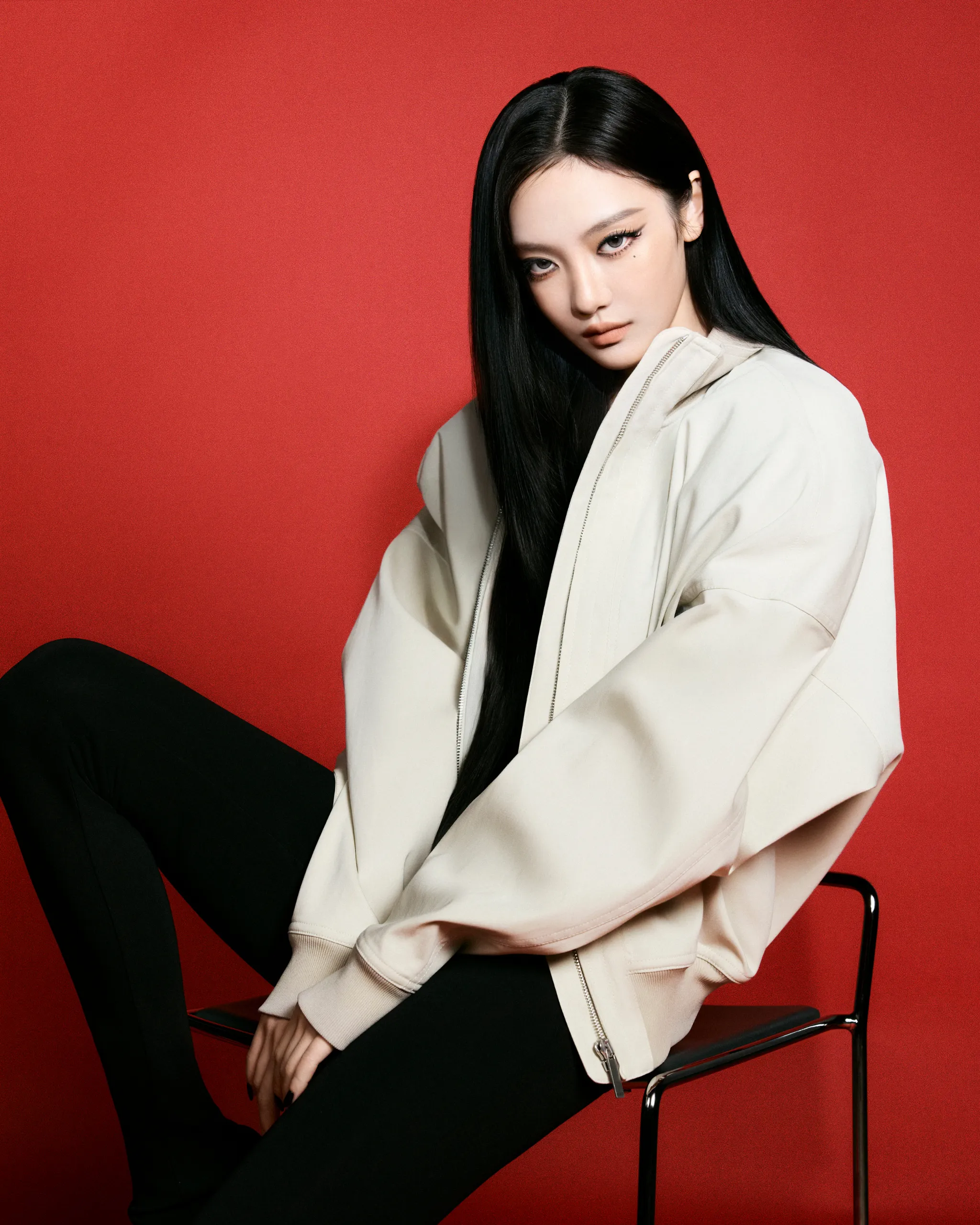 Portrait of a woman with long black hair, wearing a beige jacket and black pants, posed against a solid red backdrop.