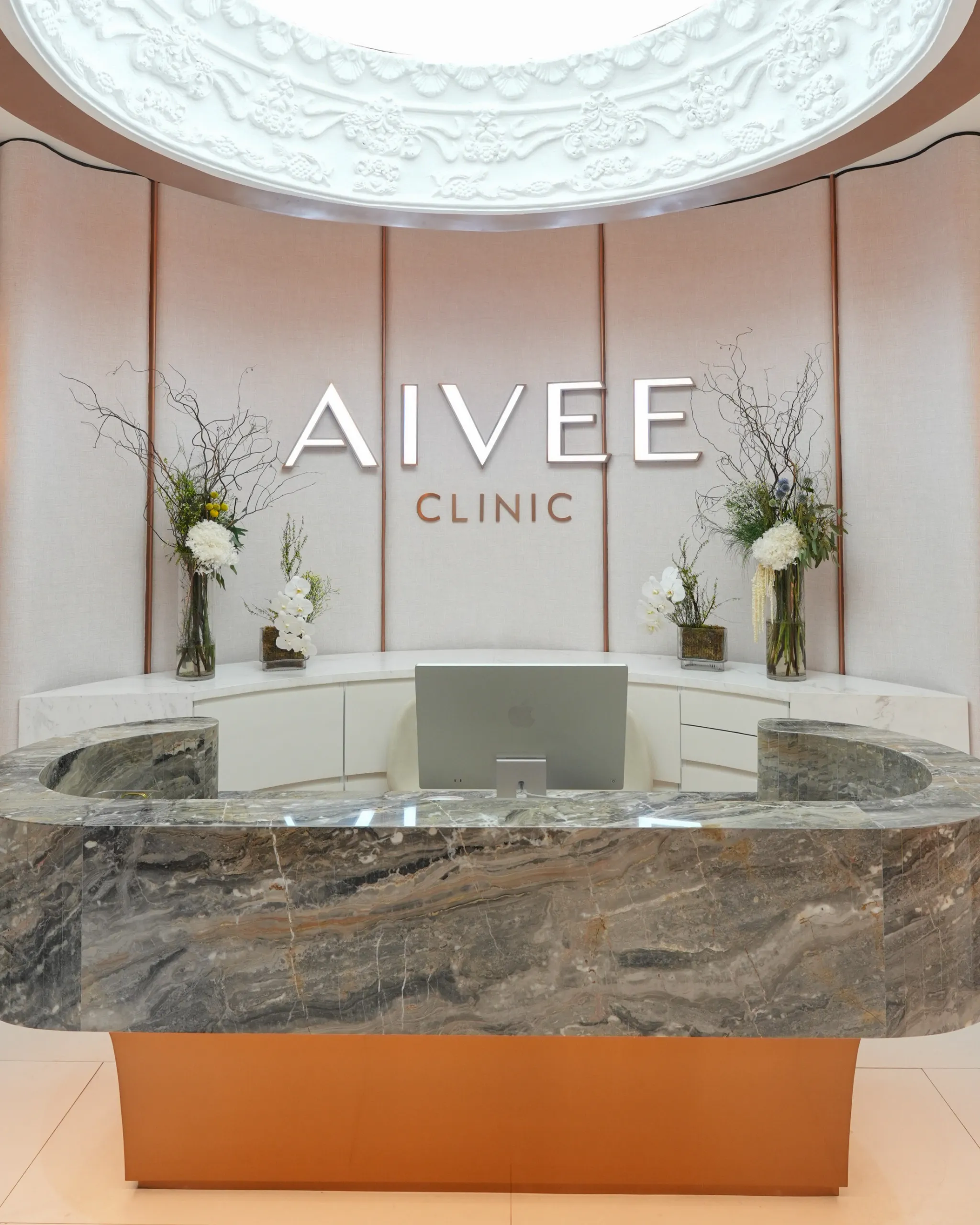 Reception area featuring a curved marble desk and light pink fabric panels behind the wall, with large 'AIVEE CLINIC' signage and decorative floral arrangements on either side