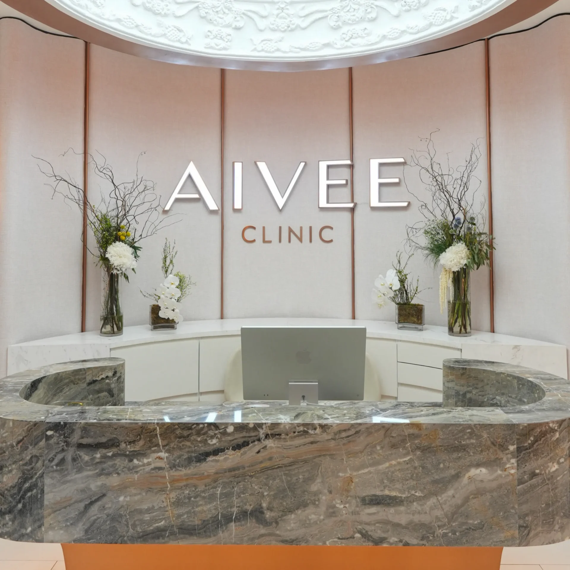 Modern clinic reception area with a curved white marble counter and large back panel displaying 'ALVE' and 'CLINIC', flanked by tall glass vases of white flowers and branches.