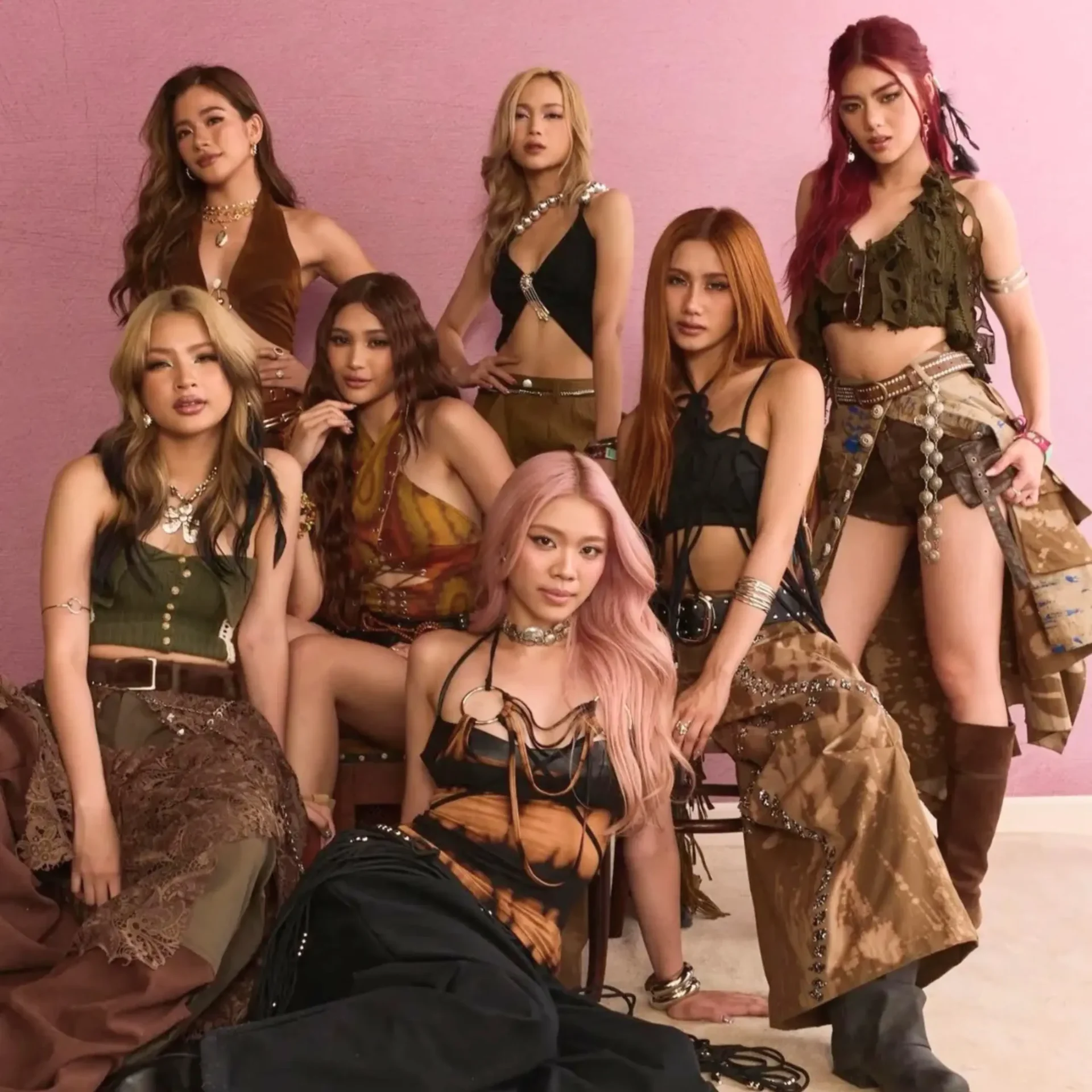 Group portrait of seven women in earthy-toned outfits posing against a pink wall.