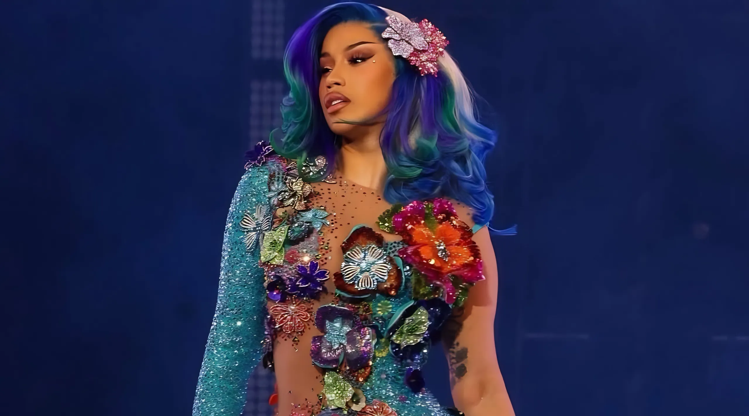 Performer with vibrant multicolor hair and a flower-adorned sequined outfit on stage against a blue background.