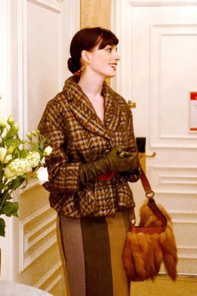 Woman in a brown check coat with gloves, standing in a light-colored hallway, holding a brown fur handbag and smiling.