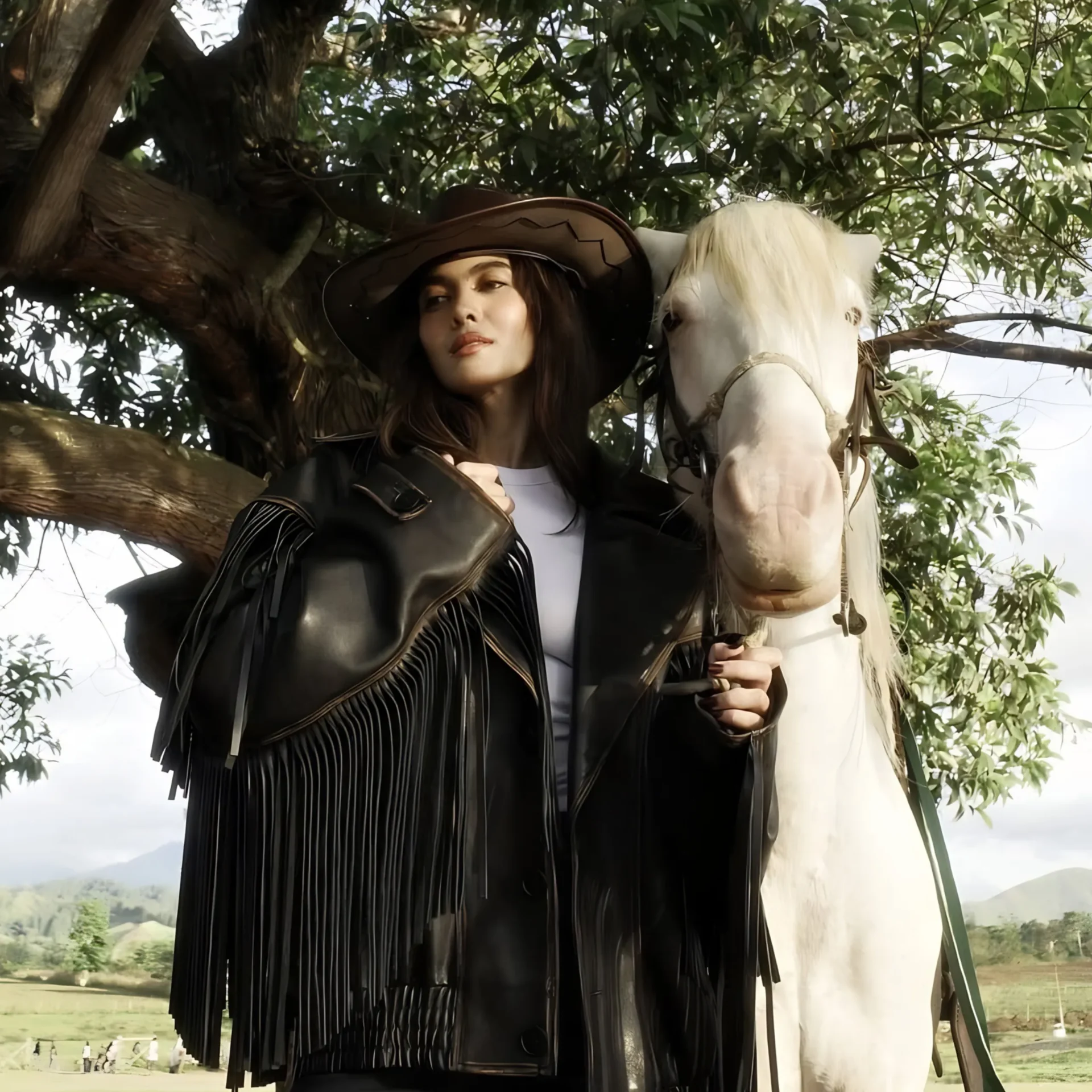 Woman in a wide-brimmed hat and fringed black jacket beside a white horse in a tree-filled outdoor setting.
