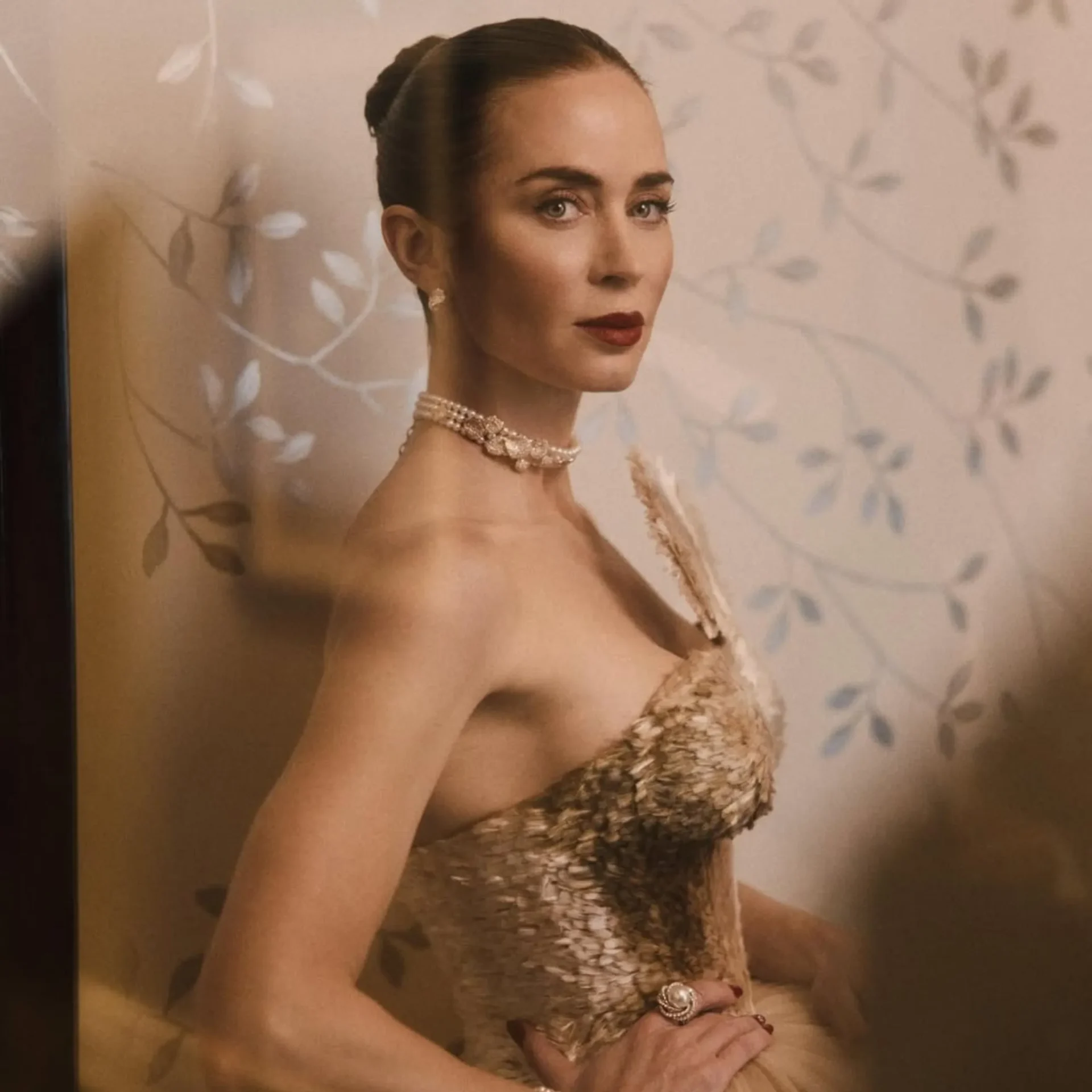 Portrait of a woman in an ornate, strapless dress and pearl necklace, arms crossed, looking to the side with a composed expression.