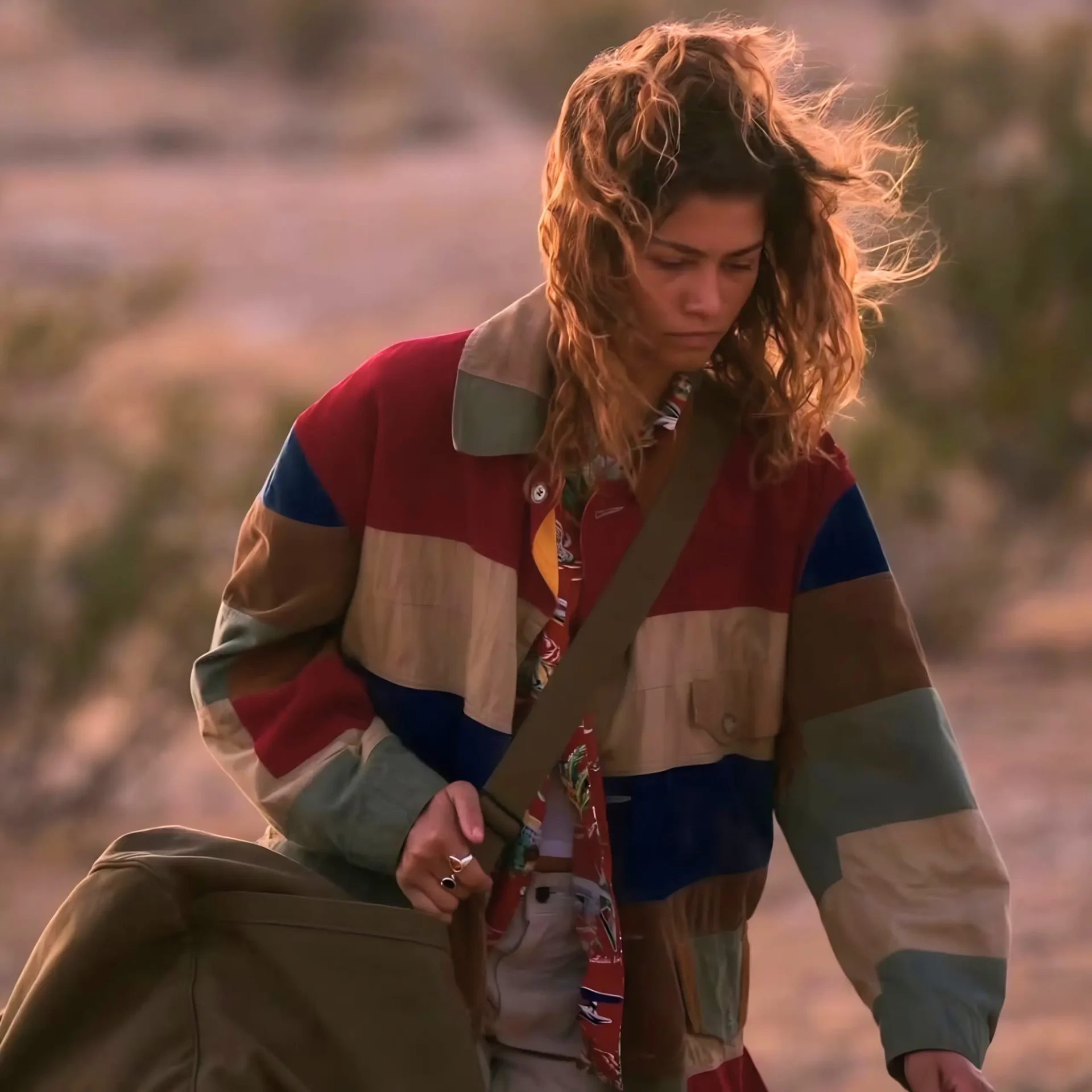 Woman with windblown hair in a multicolor striped jacket, adjusting a large tan bag outdoors.