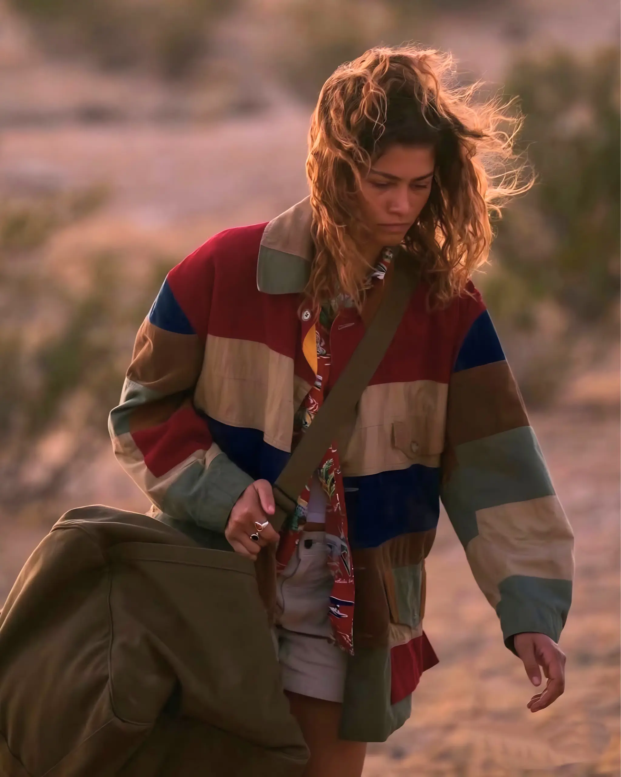Woman with wavy hair wearing a large, color-blocked jacket and a green duffel bag outdoors.