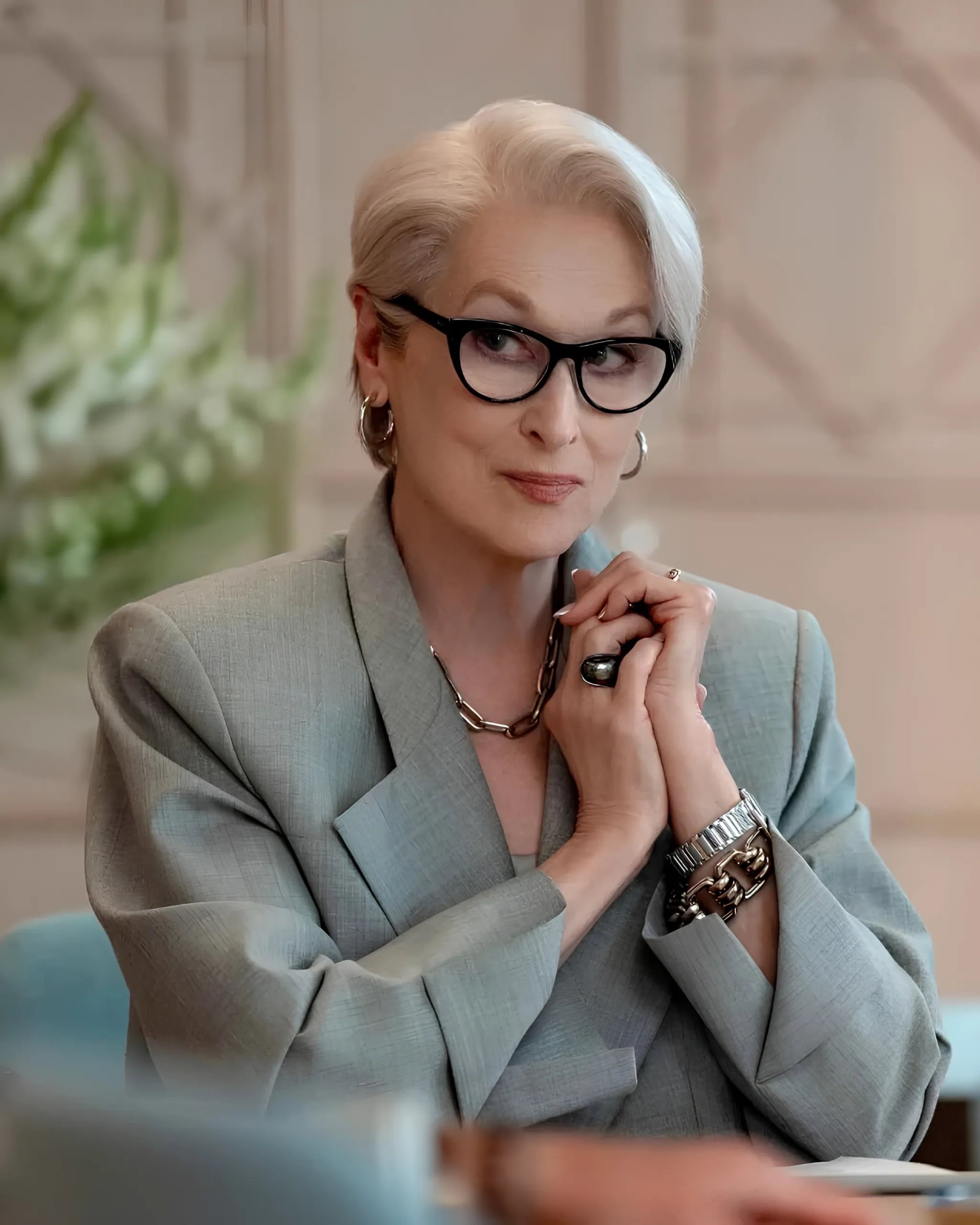 Professional woman with short blonde hair and black glasses, wearing a gray blazer, resting her chin on clasped hands.