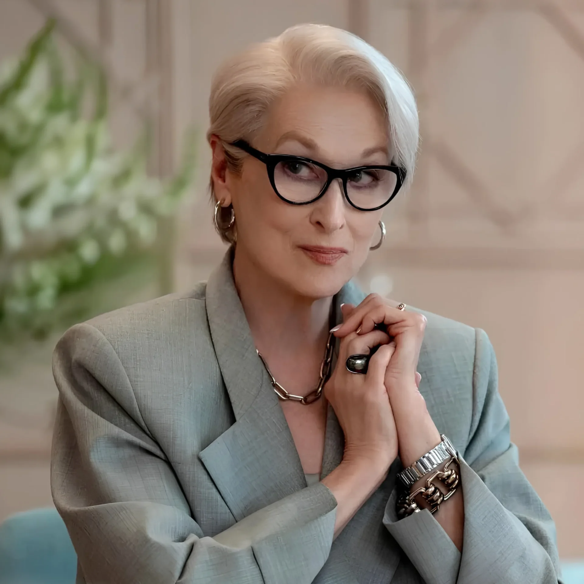Woman with short silver hair and black glasses, wearing a gray blazer, resting her chin on clasped hands in a thoughtful pose.