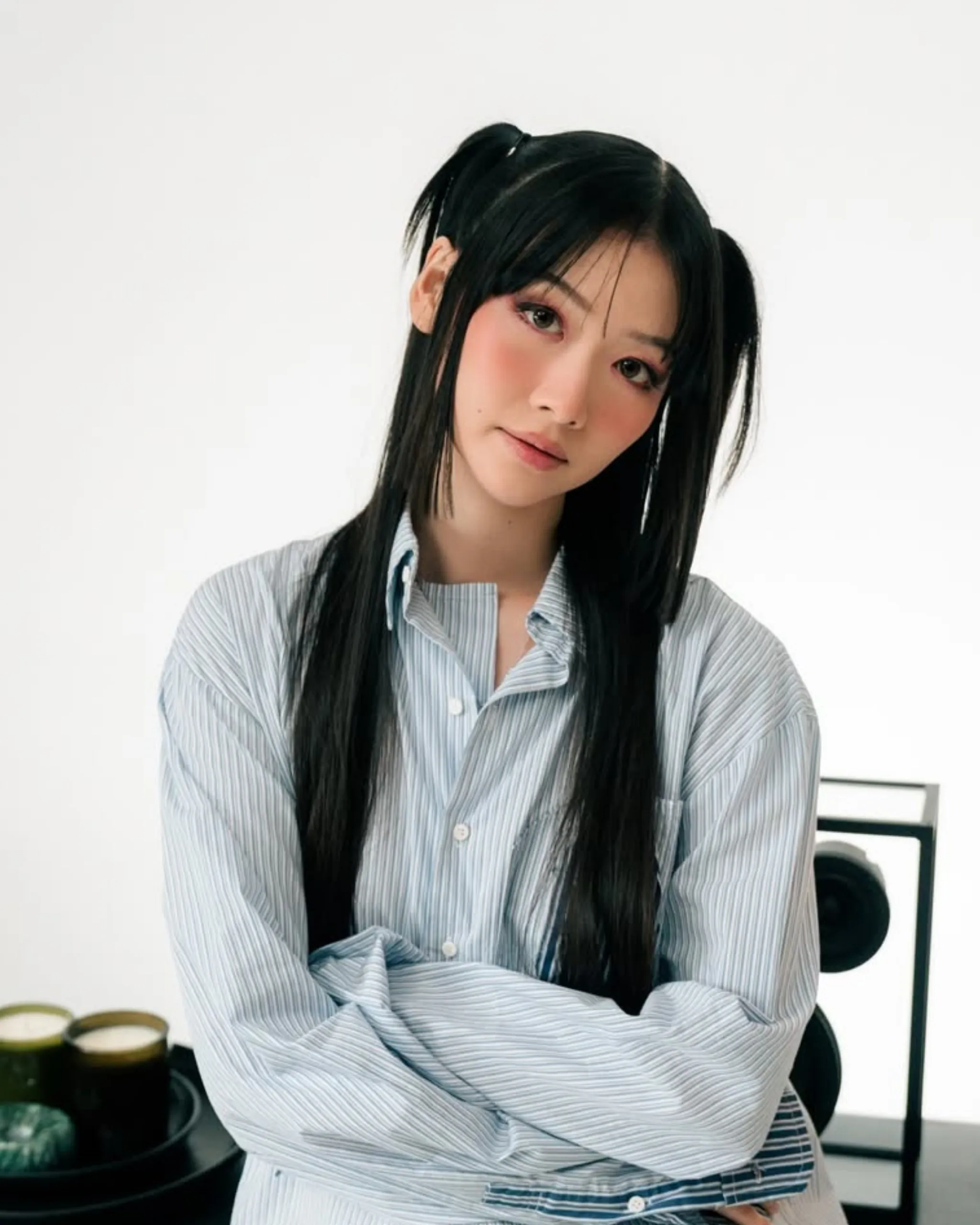 Young woman with long black hair in a blue striped shirt sits with arms crossed, looking at the camera against a white background.