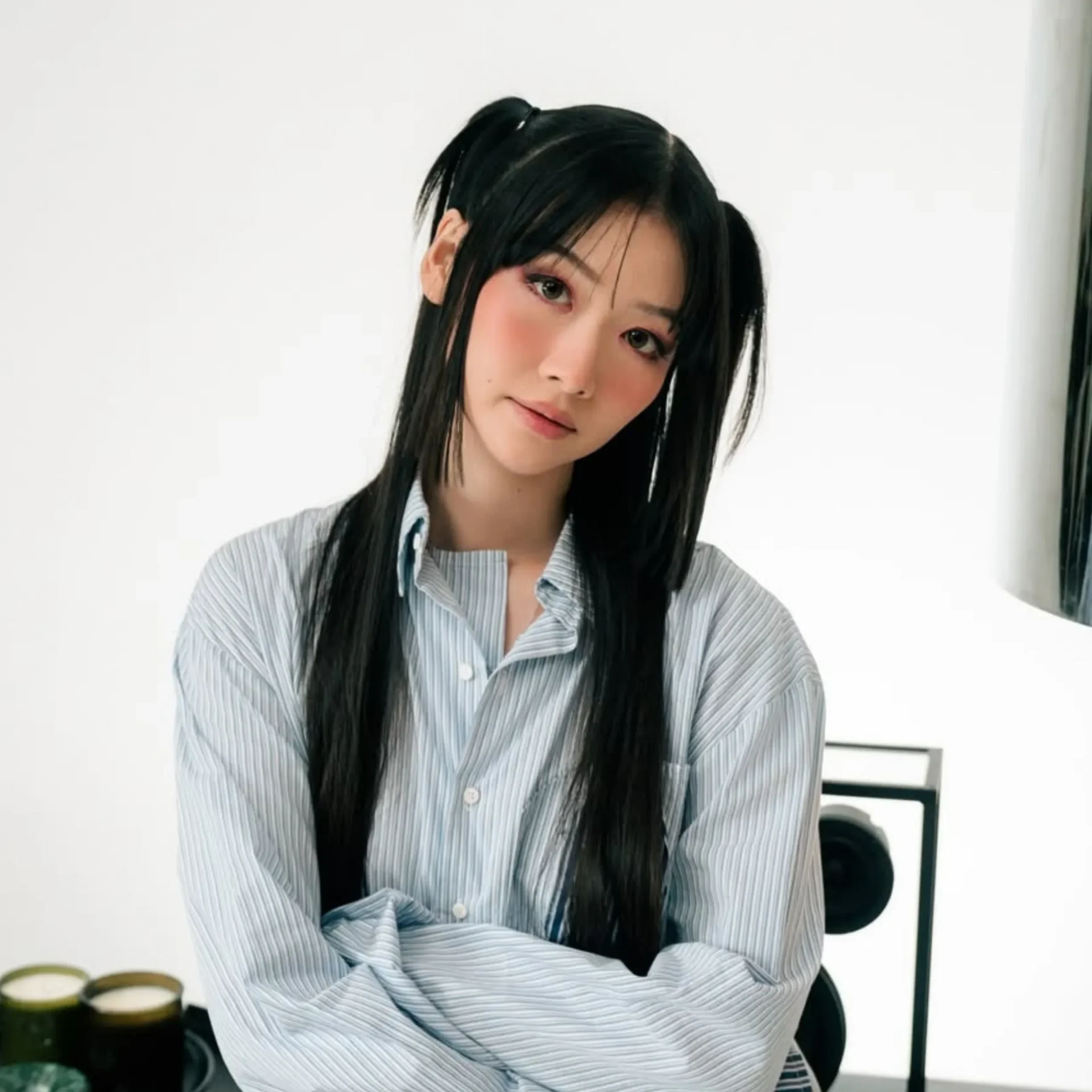 Young woman with long black hair wearing a striped button-up shirt, arms crossed, in a bright indoor setting.