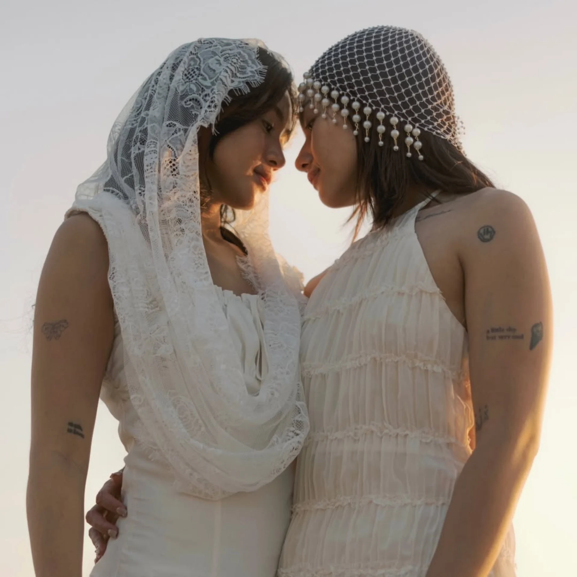 Two women in white lace dresses with veils lean foreheads together in a tender moment of affection.
