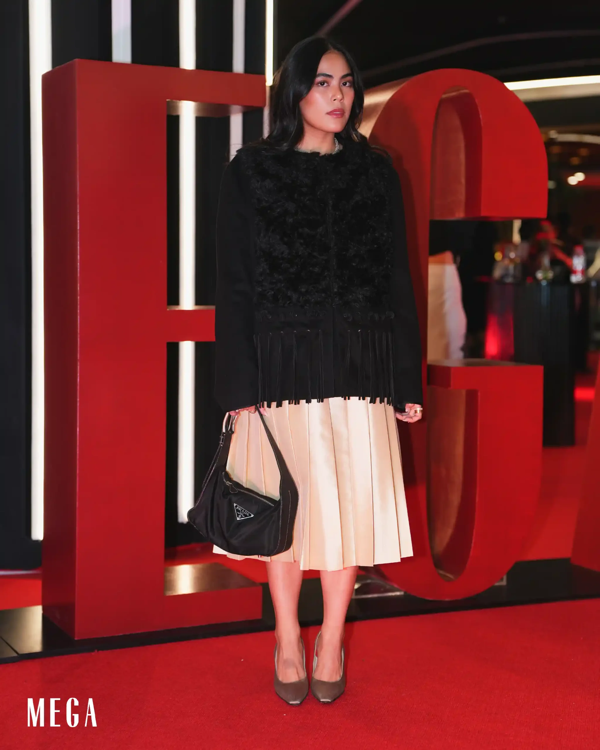 Woman posing on a red carpet in a black textured top and beige pleated skirt, holding a black handbag, with a red modern backdrop and MEGA logo visible.