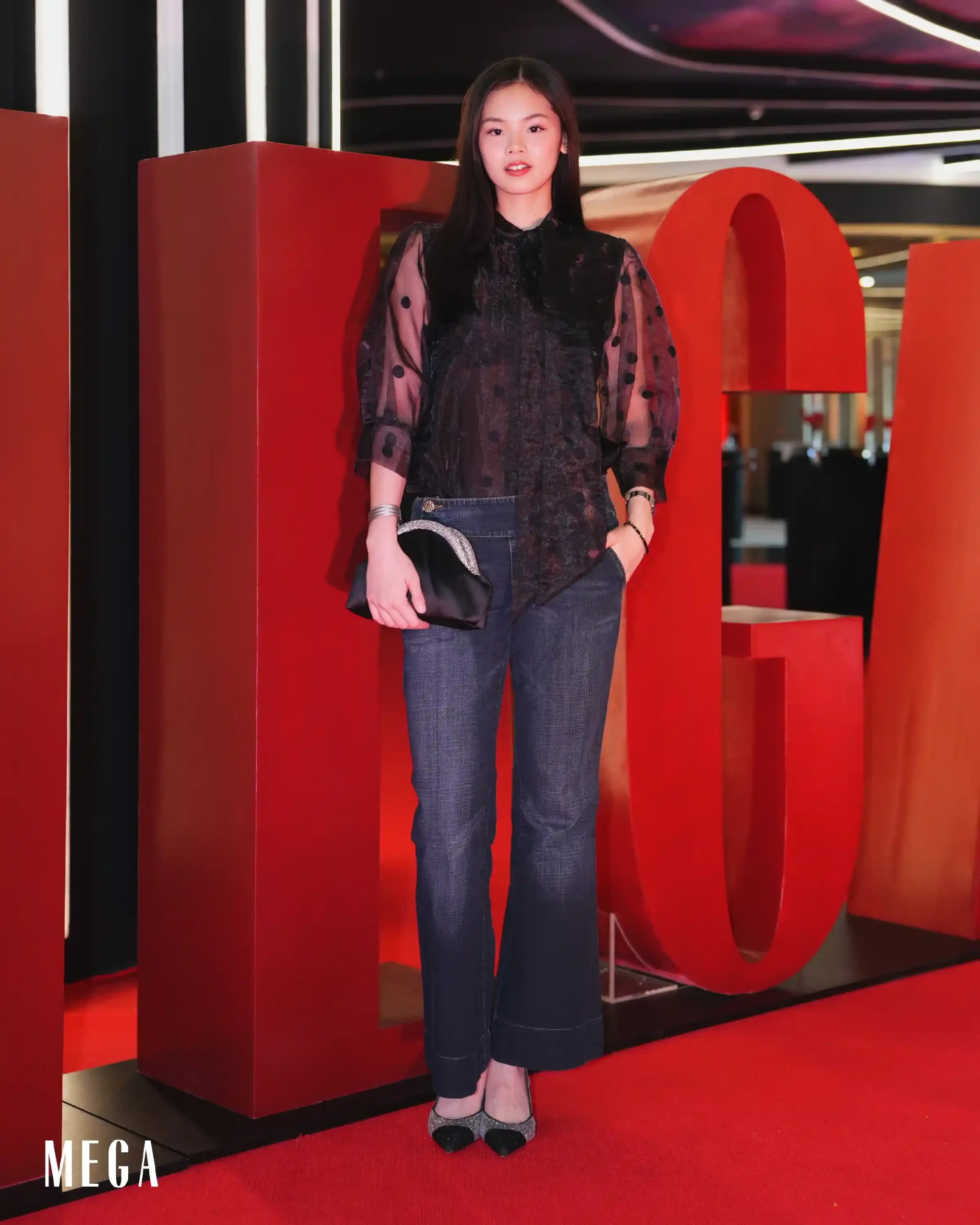 Woman standing in front of large red decorative letters at an event, wearing a black lace blouse and jeans, posing for the camera.