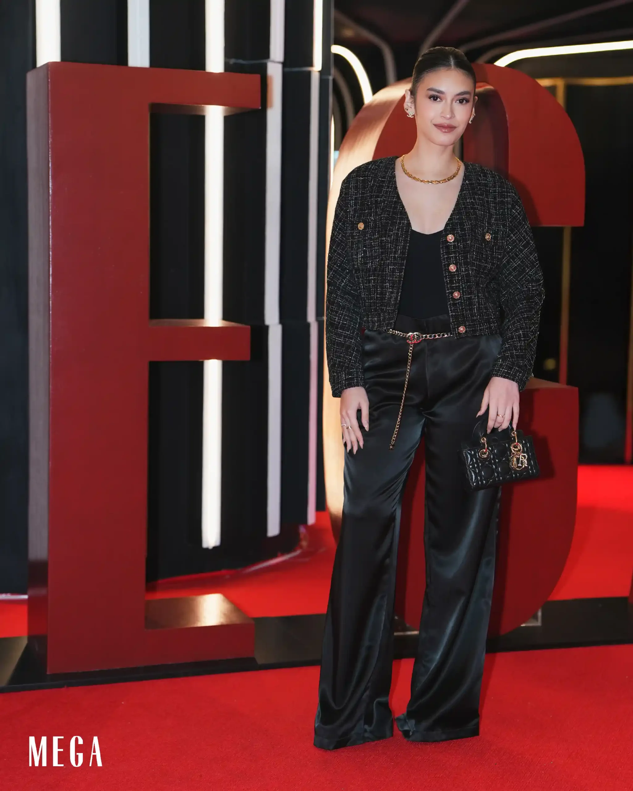 Woman in a black tweed jacket and satin pants posing on a red carpet with large red MEGA letters in the background.