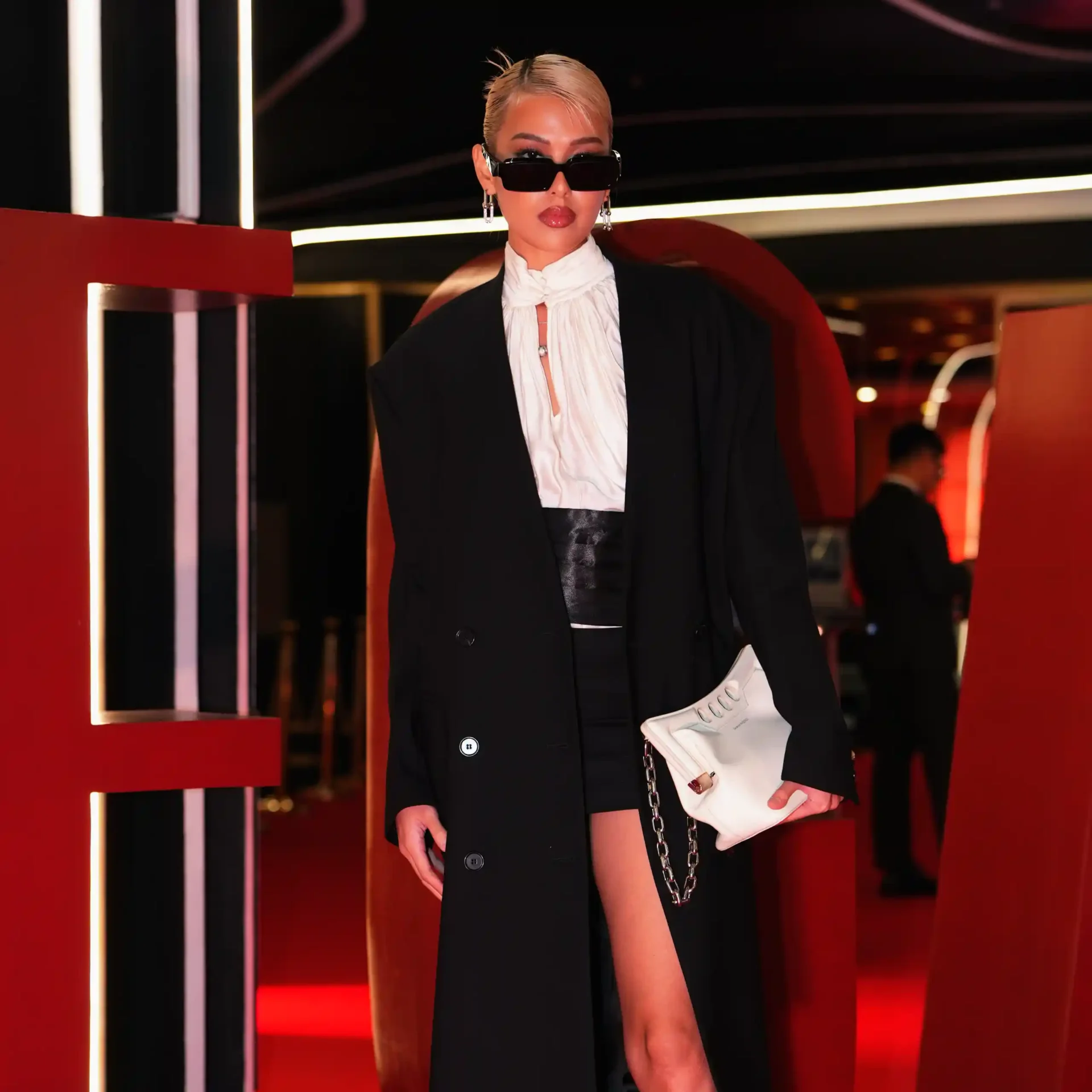 Person in a black blazer over a white ruffled blouse, wearing oversized black sunglasses, with a white handbag, in a red-lit interior.