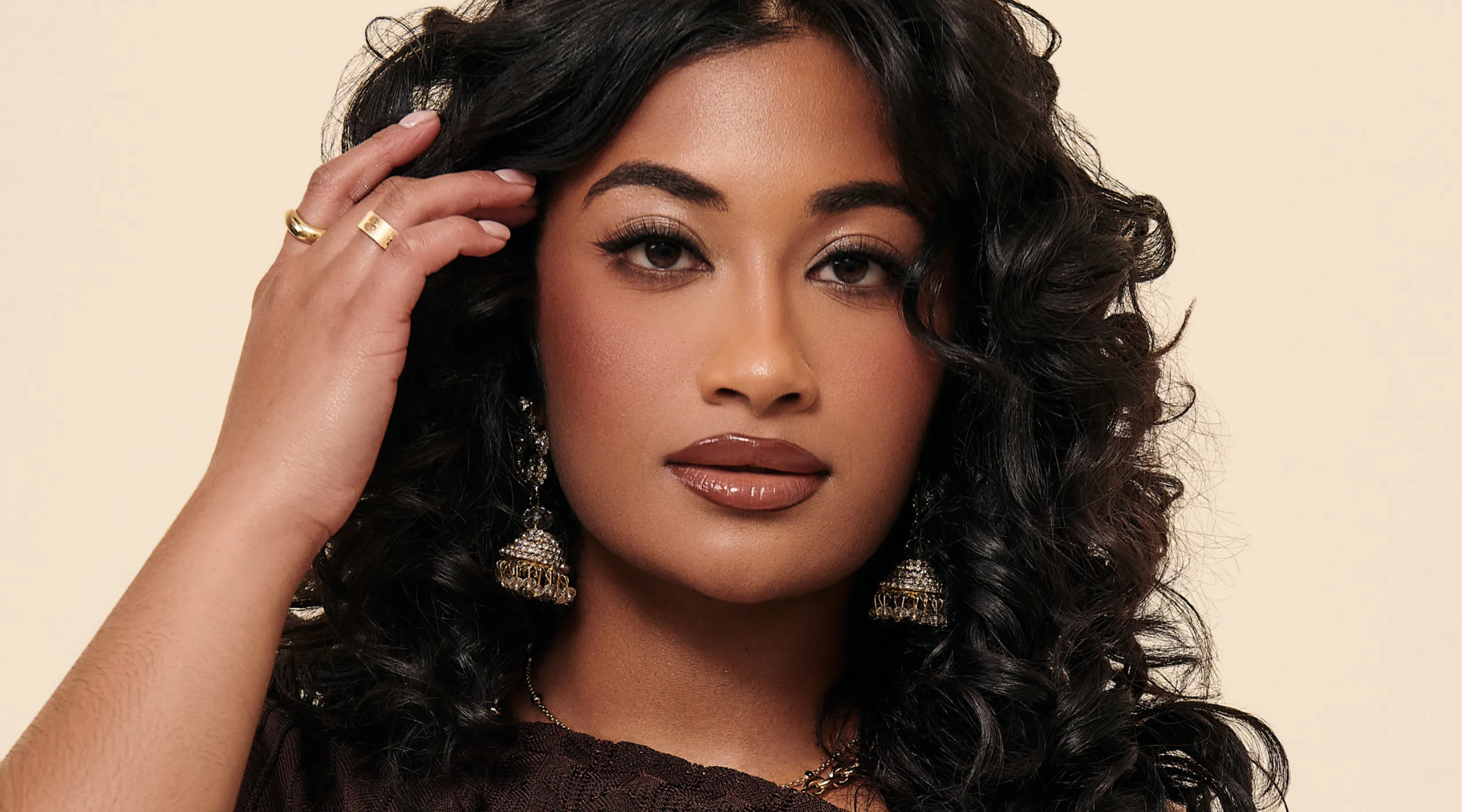 Close-up portrait of a woman with dark, wavy hair, wearing gold earrings and ring, touching her hair near her temple.