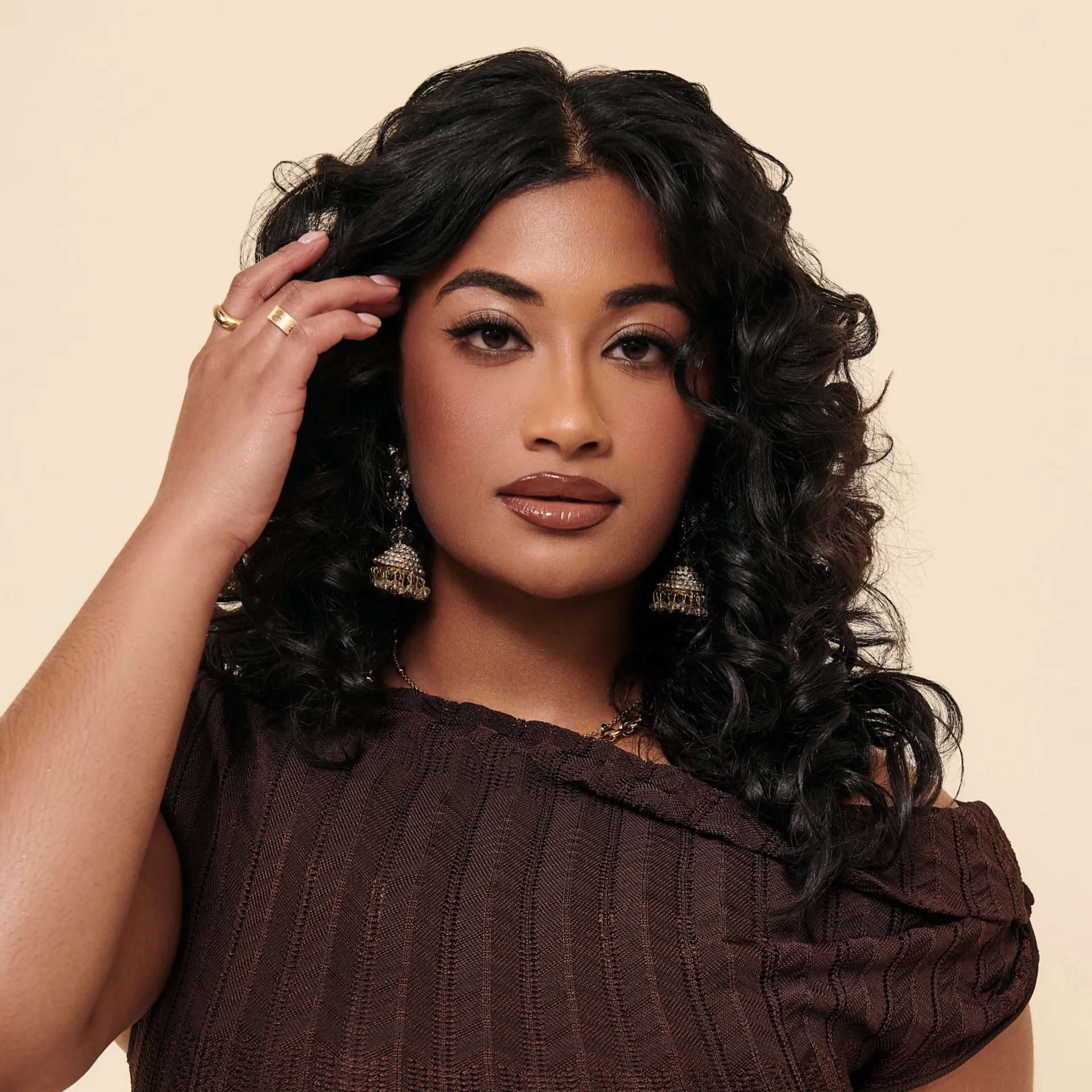 Close-up portrait of a woman with dark wavy hair/touched hair, wearing gold jewelry and a brown textured top, neutral beige background.