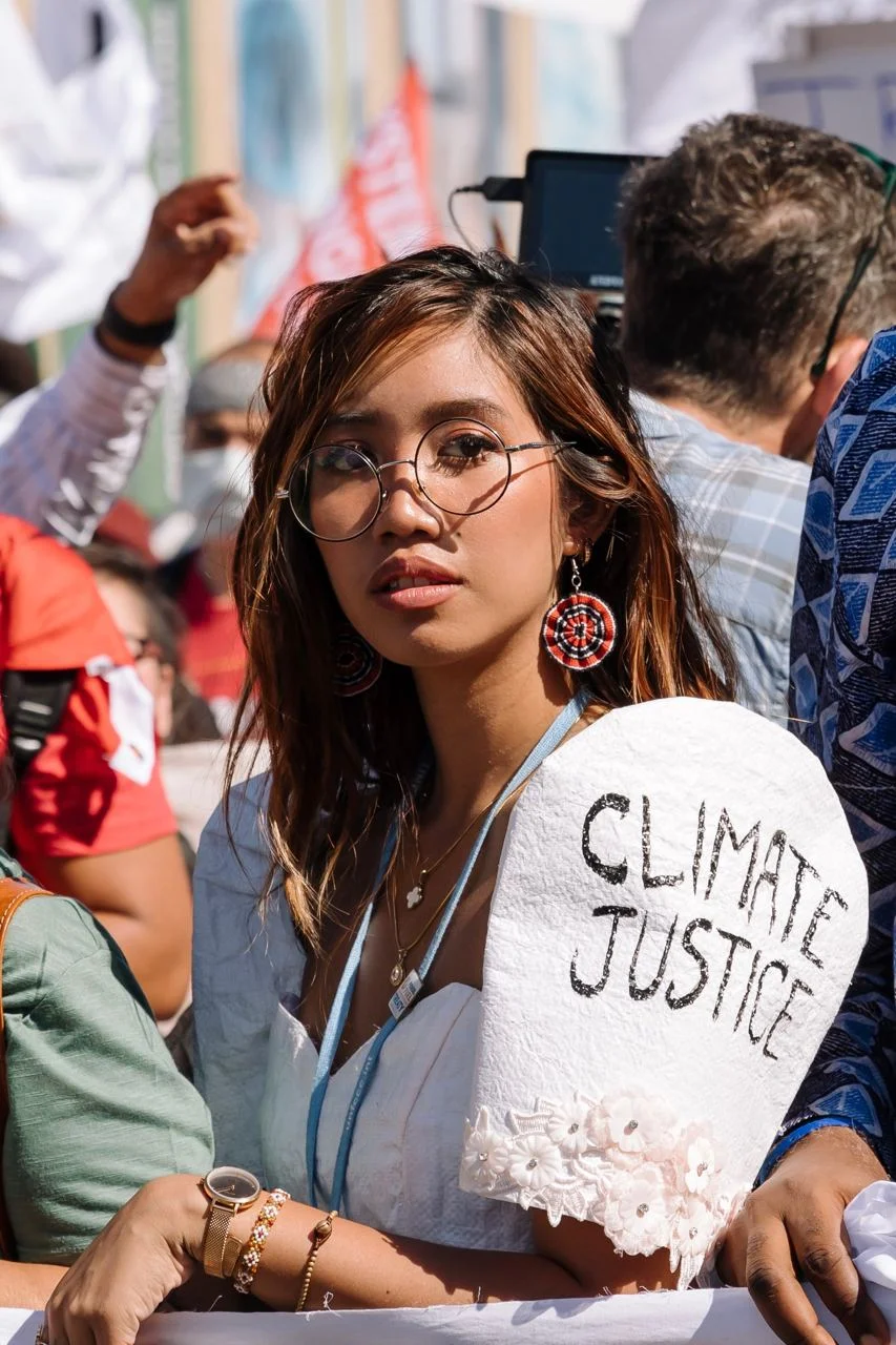 Young woman with round glasses at a climate protest holding a 'CLIMATE JUSTICE' sign. She wears white and large red earrings.