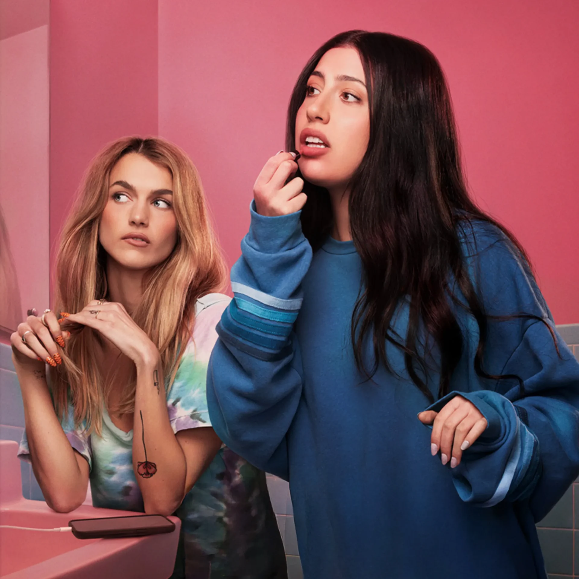 Two women in a pink-tinted bathroom; one applies lipstick while the other watches nearby at the sink,”