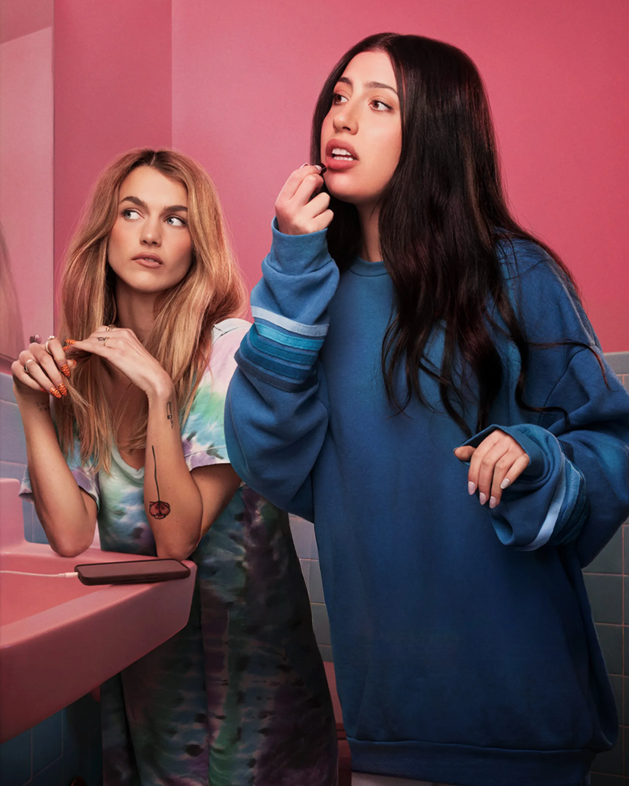 Two young women in a pink bathroom; one applies lipstick while the other watches from the side.