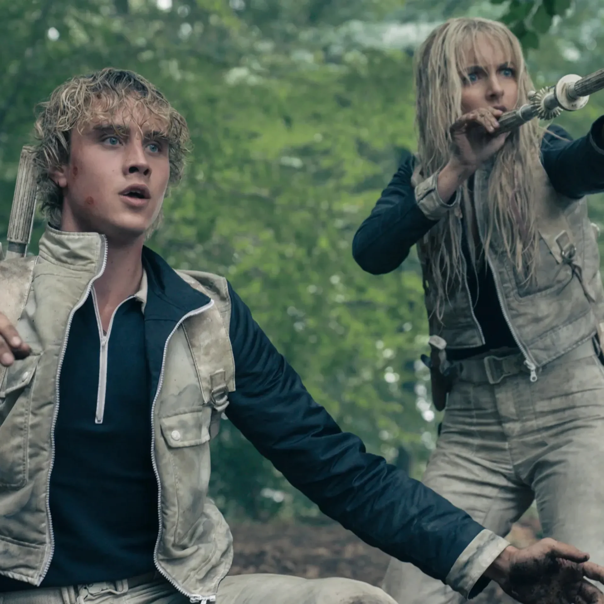 Two people in muddy survival jackets in a forest; the woman on the right holds a long, gear-tedged staff while the man looks upward.