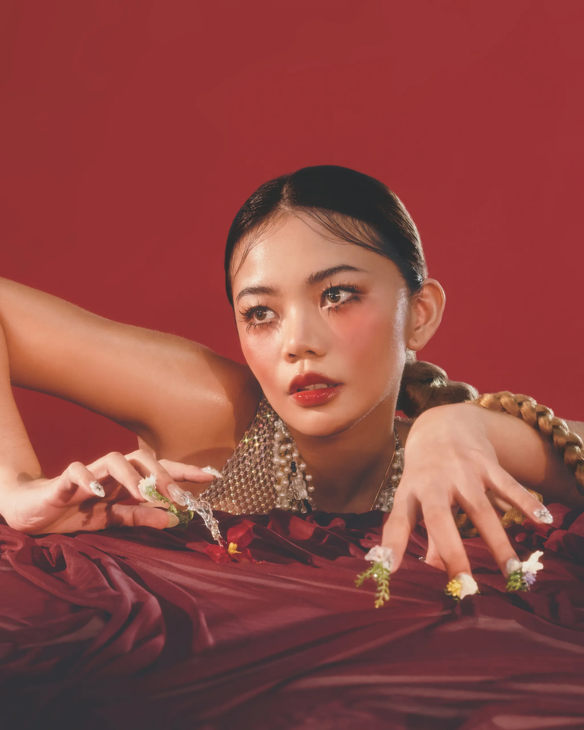 Close-up portrait of a person with braided hair and a beaded necklace against a solid red background, with hands and small flowers in the foreground.”,