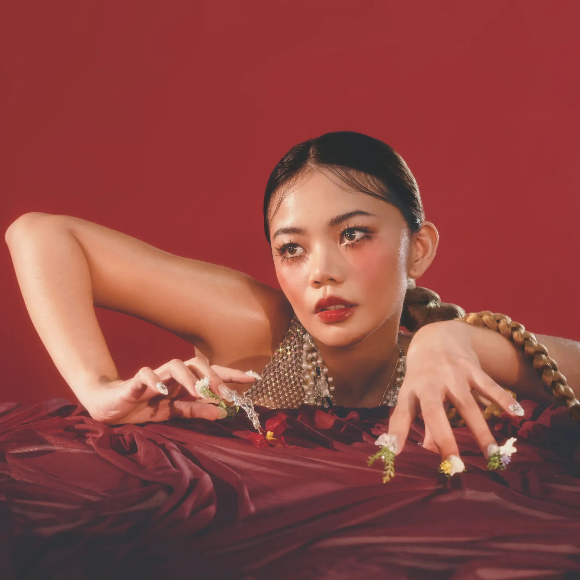 Close-up portrait of a person with braided dark hair, red lipstick, and a jeweled necklace against a red backdrop.