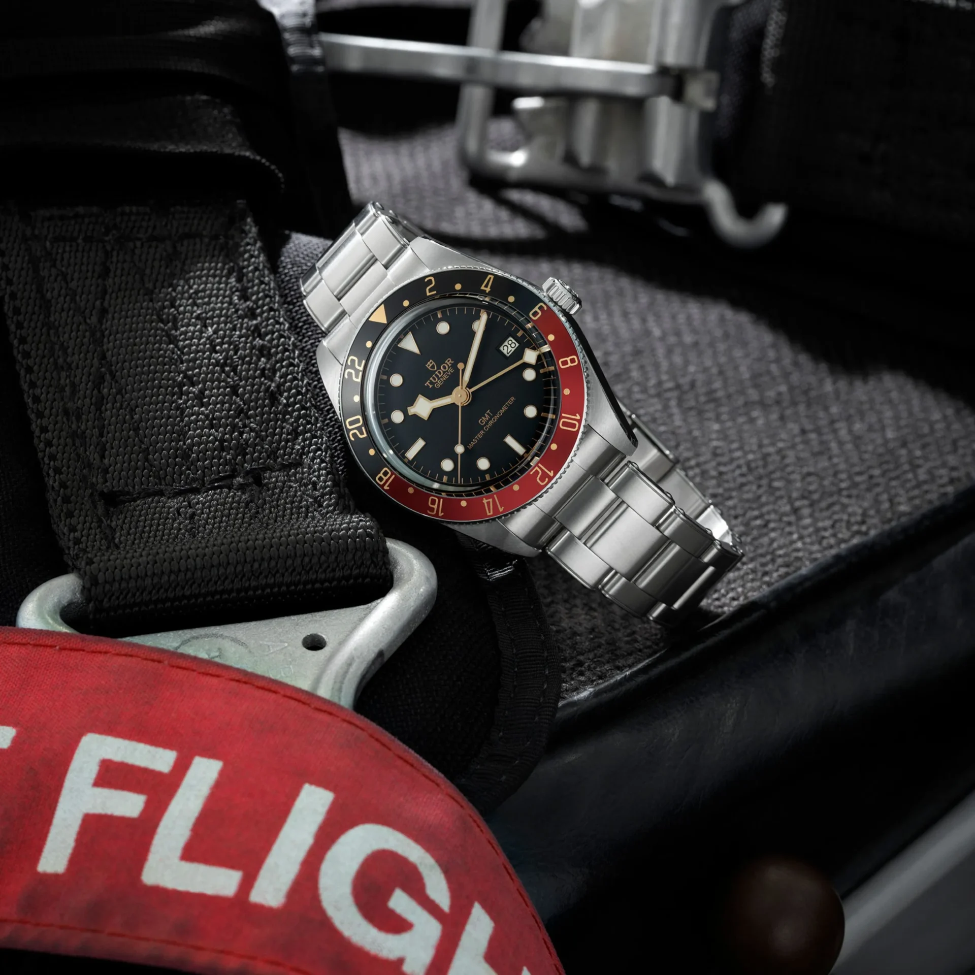 Stainless steel Tudor GMT wristwatch with black dial and red/black bezel on tactical gear.