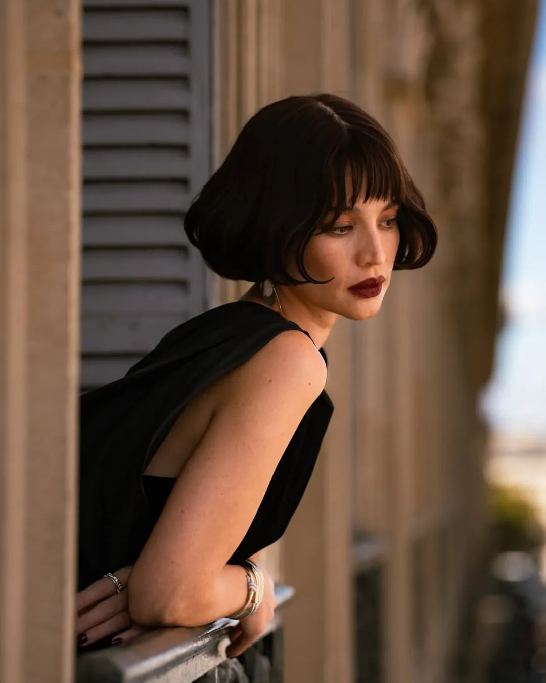 Woman with short dark hair leaning on a railing, wearing a black sleeveless dress, gazing thoughtfully downward outdoors.