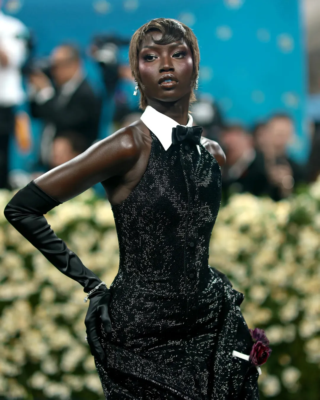 Model on a red carpet wearing a sparkly black tuxedo with a white wing collar, black bow tie, and elbow-length gloves, holding a flower corsage.