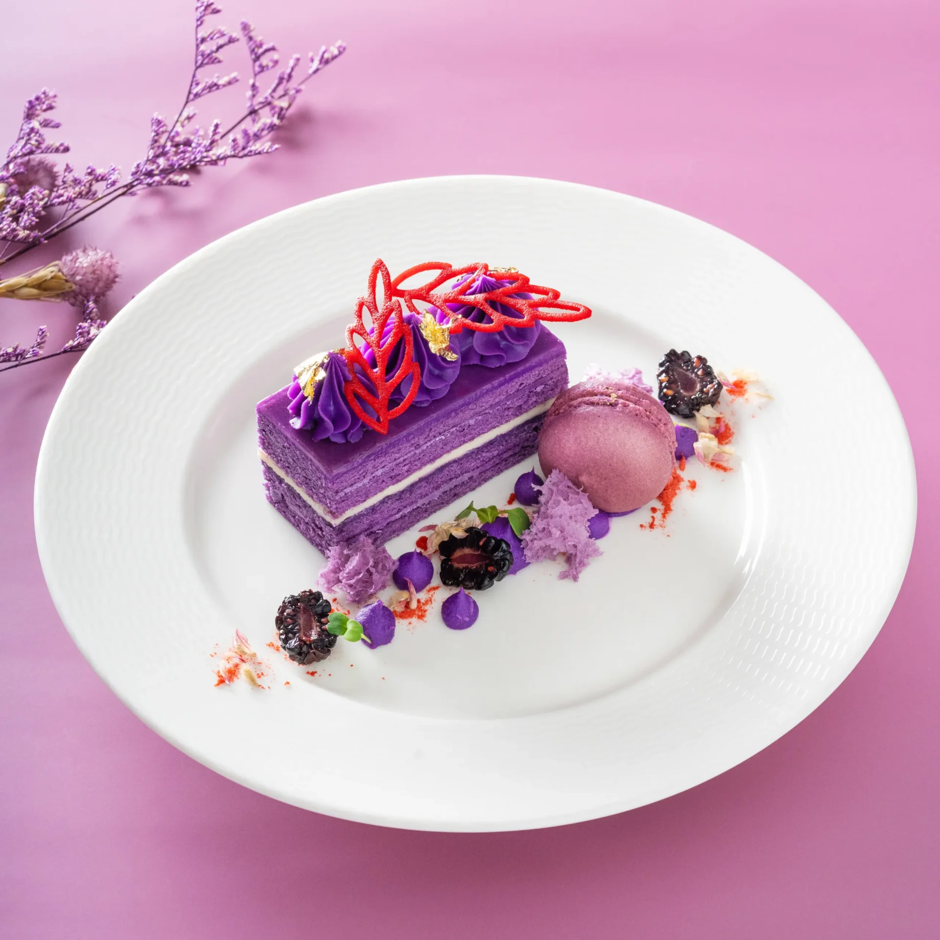 Purple layered cake slice with purple frosting and red lattice topping on a white textured plate against a pink background.